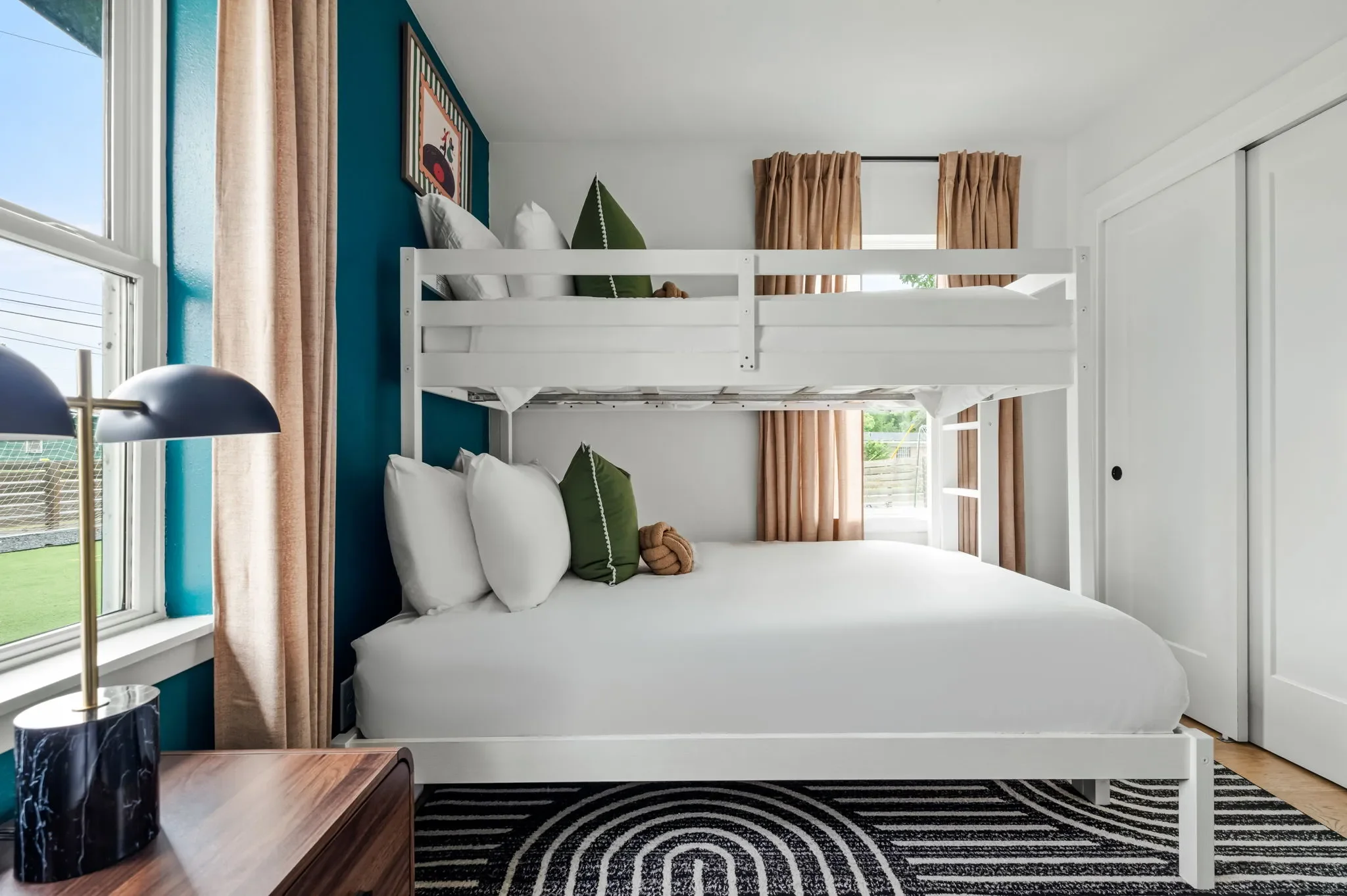 Airbnb bunk room design detail with white bunk beds, teal accent wall, green pillows, navy task lamp, and graphic rug — Austin TX.