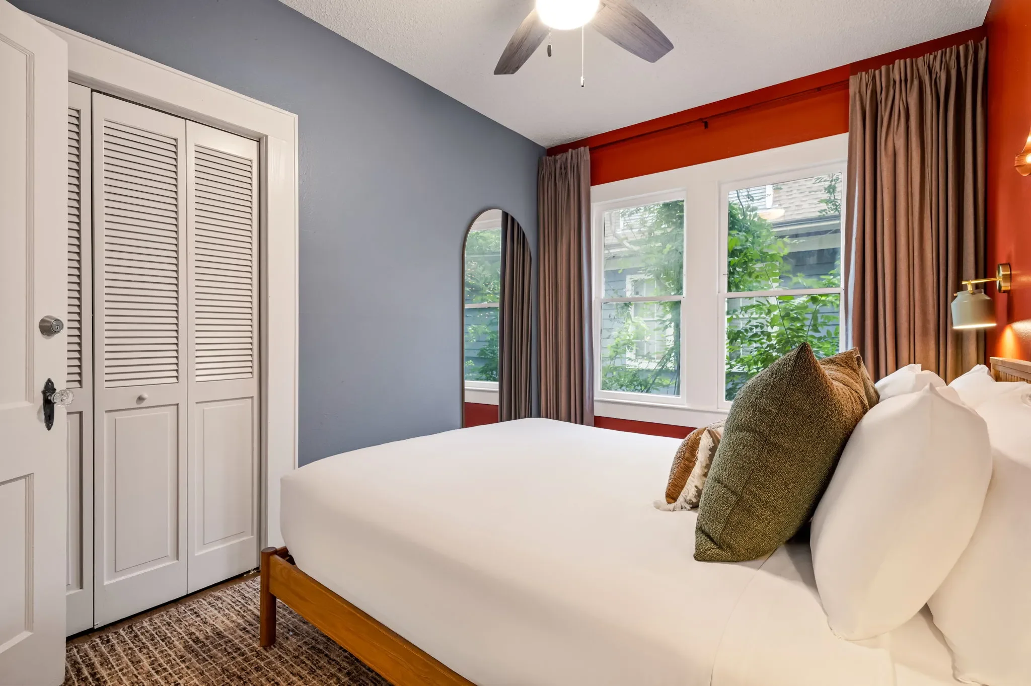 Airbnb bedroom design with orange accent wall, blue-gray walls, wood bed frame, green pillows, and arched mirror — Austin TX.