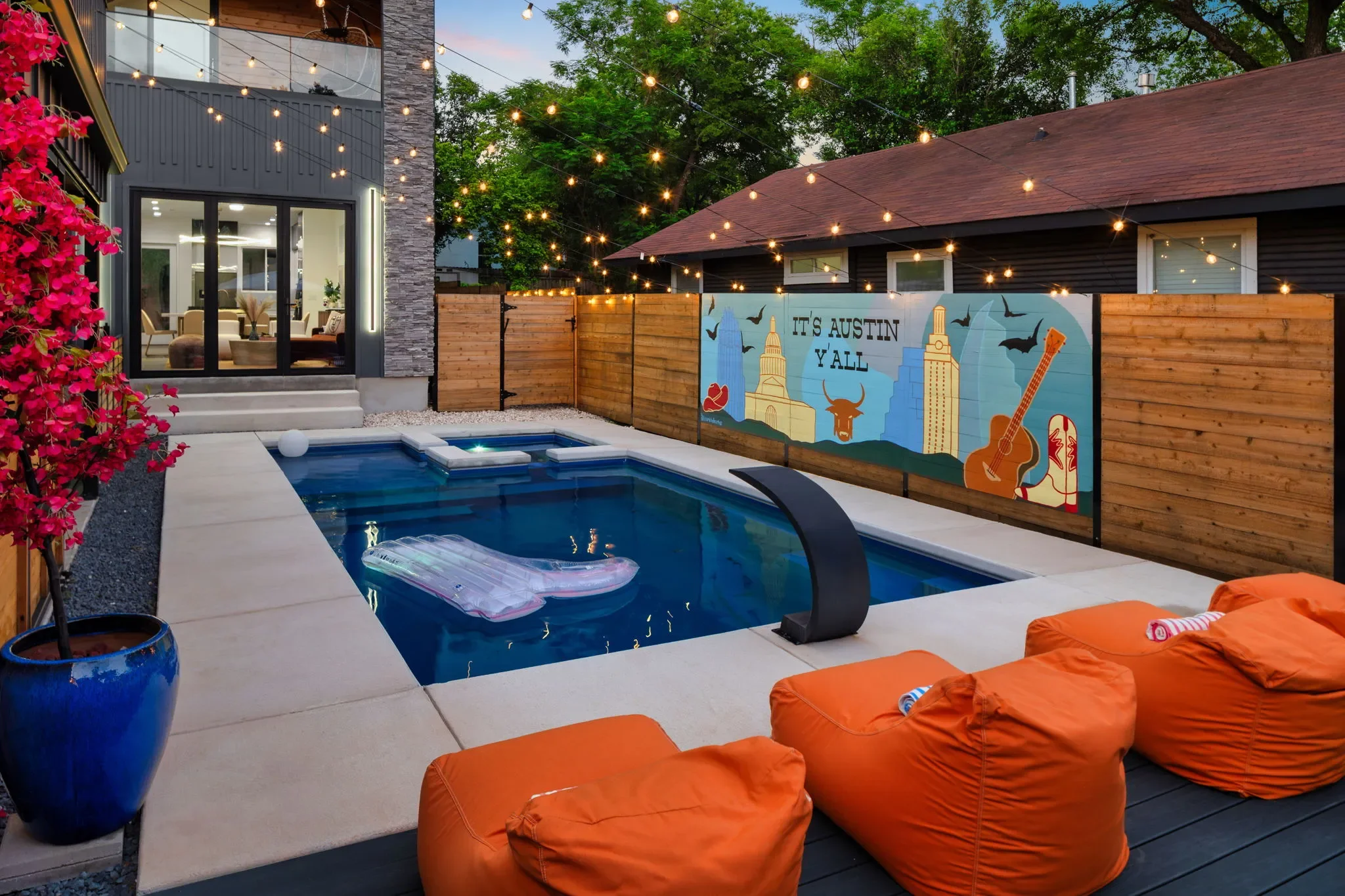 Outdoor backyard with a large pool and hot tub, wall mural reading “It’s Austin Y’all,” and orange lounge chairs.