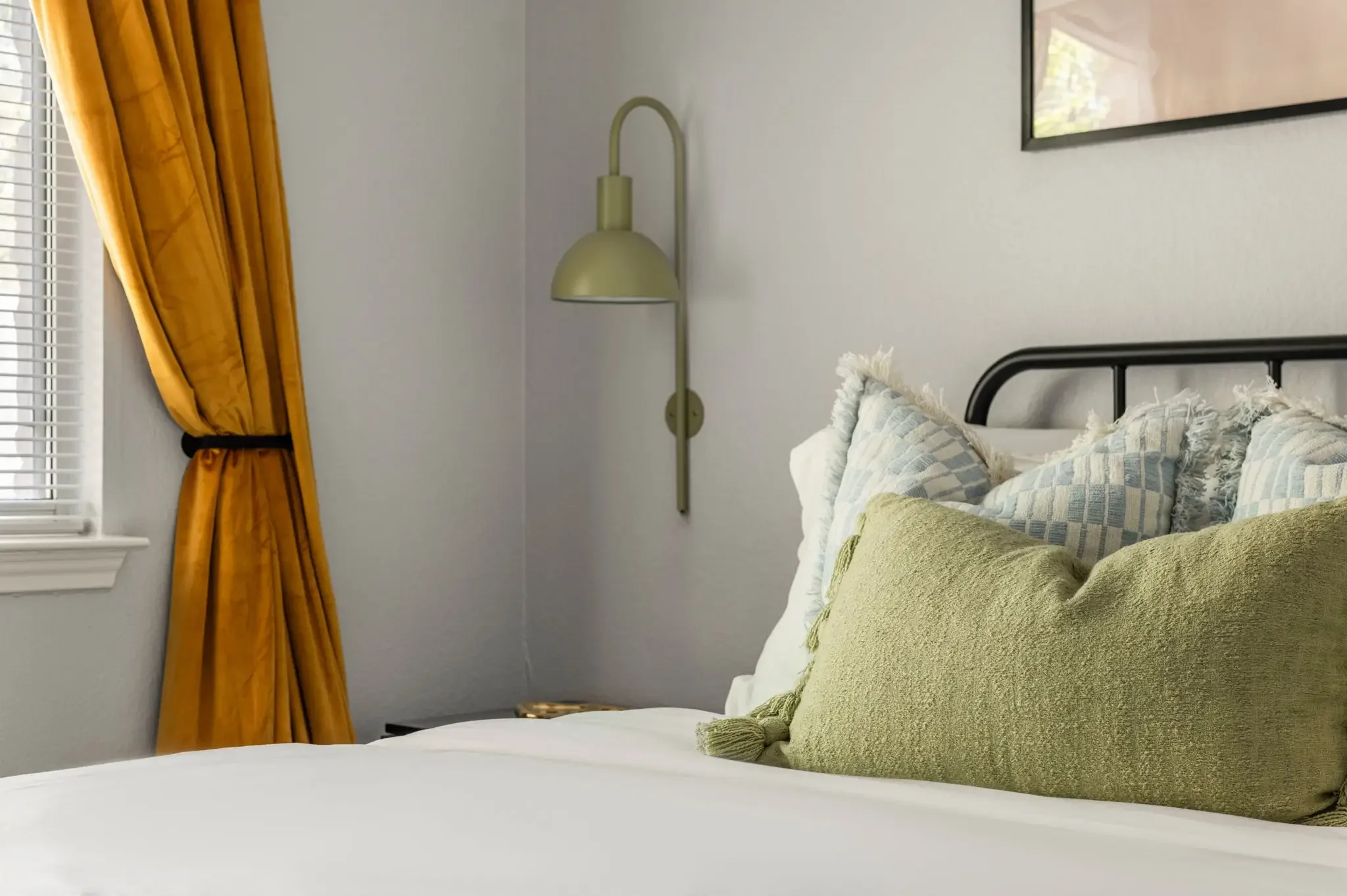 Airbnb bedroom styling detail with gold curtains, black iron headboard, sage green sconce, and layered pillows — Austin TX.