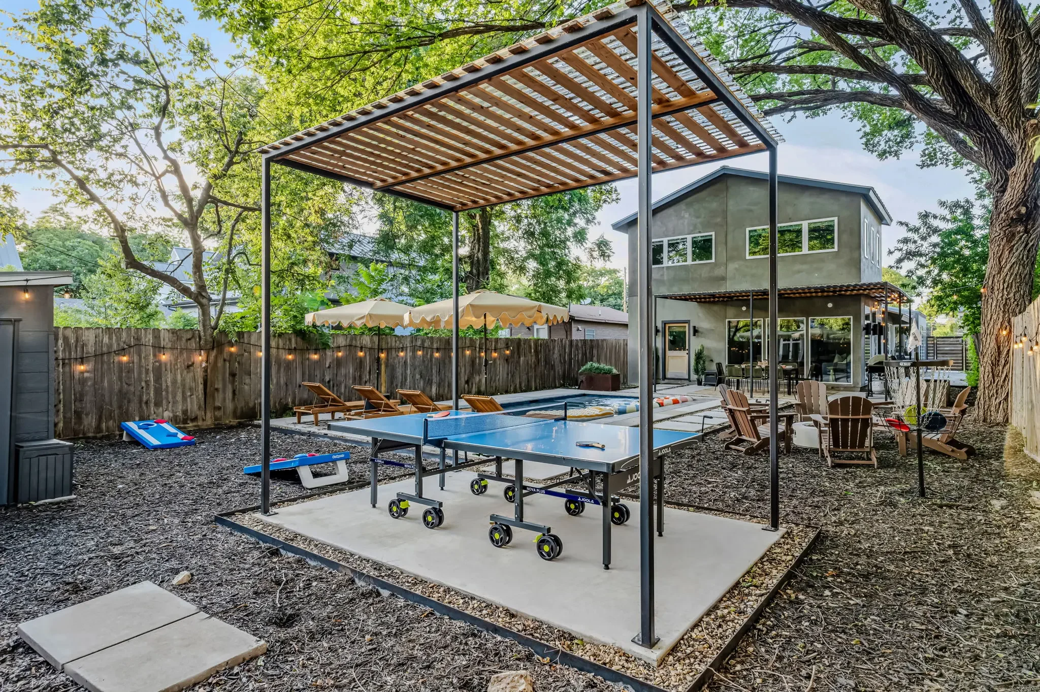 Short-term rental backyard design with ping pong table under pergola, cornhole boards, pool with scalloped umbrella, and Adirondack chairs — Austin TX.