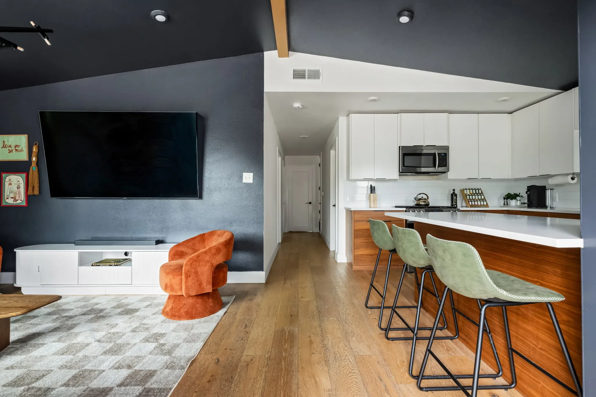 Short-term rental kitchen and living area design with white cabinets, wood island, green bar stools, large TV, and dark accent wall — Austin TX.