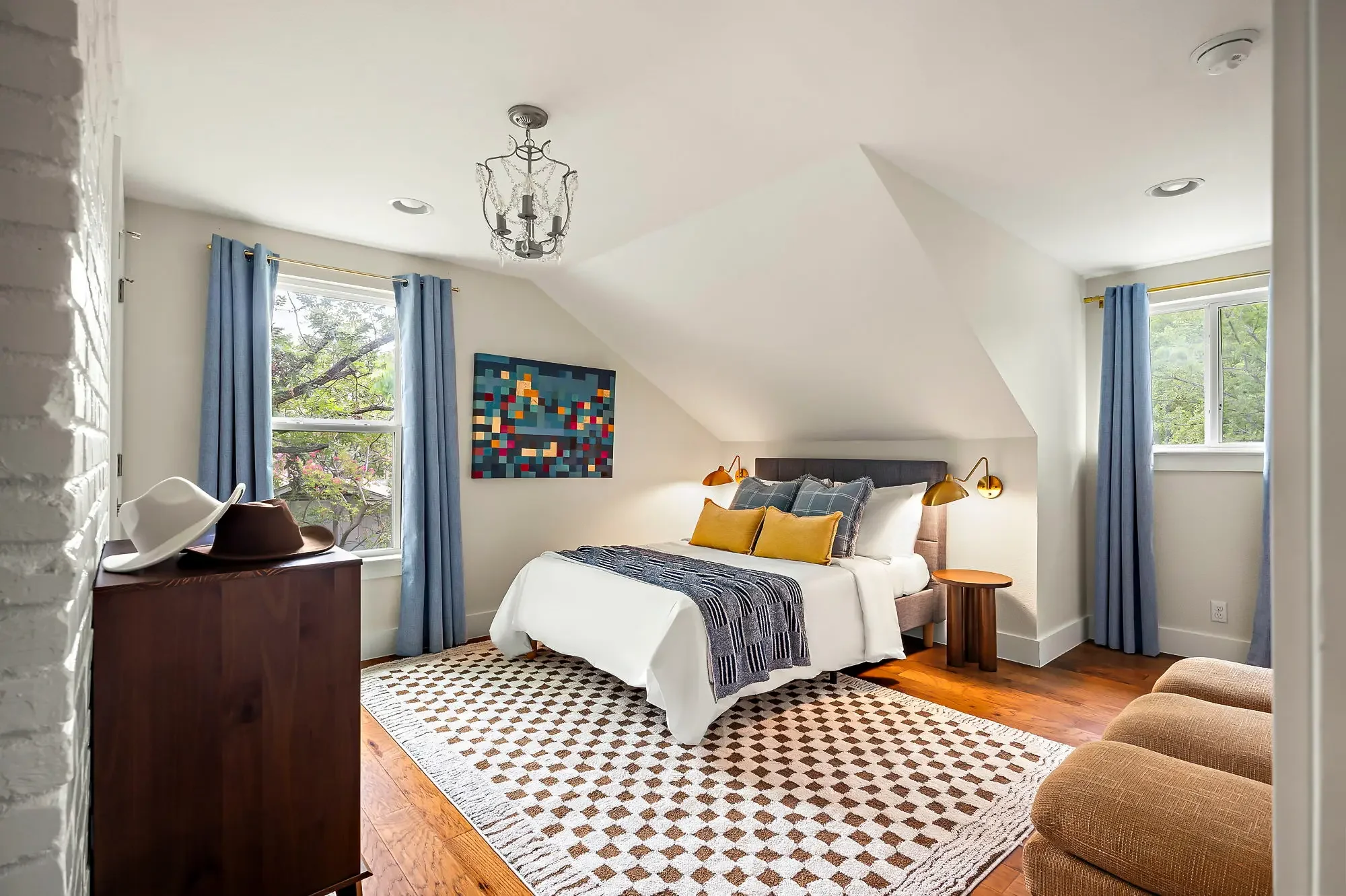 Short-term rental attic bedroom design with blue curtains, checkered rug, colorful mosaic wall art, gold sconce, and vaulted ceiling — Austin TX.