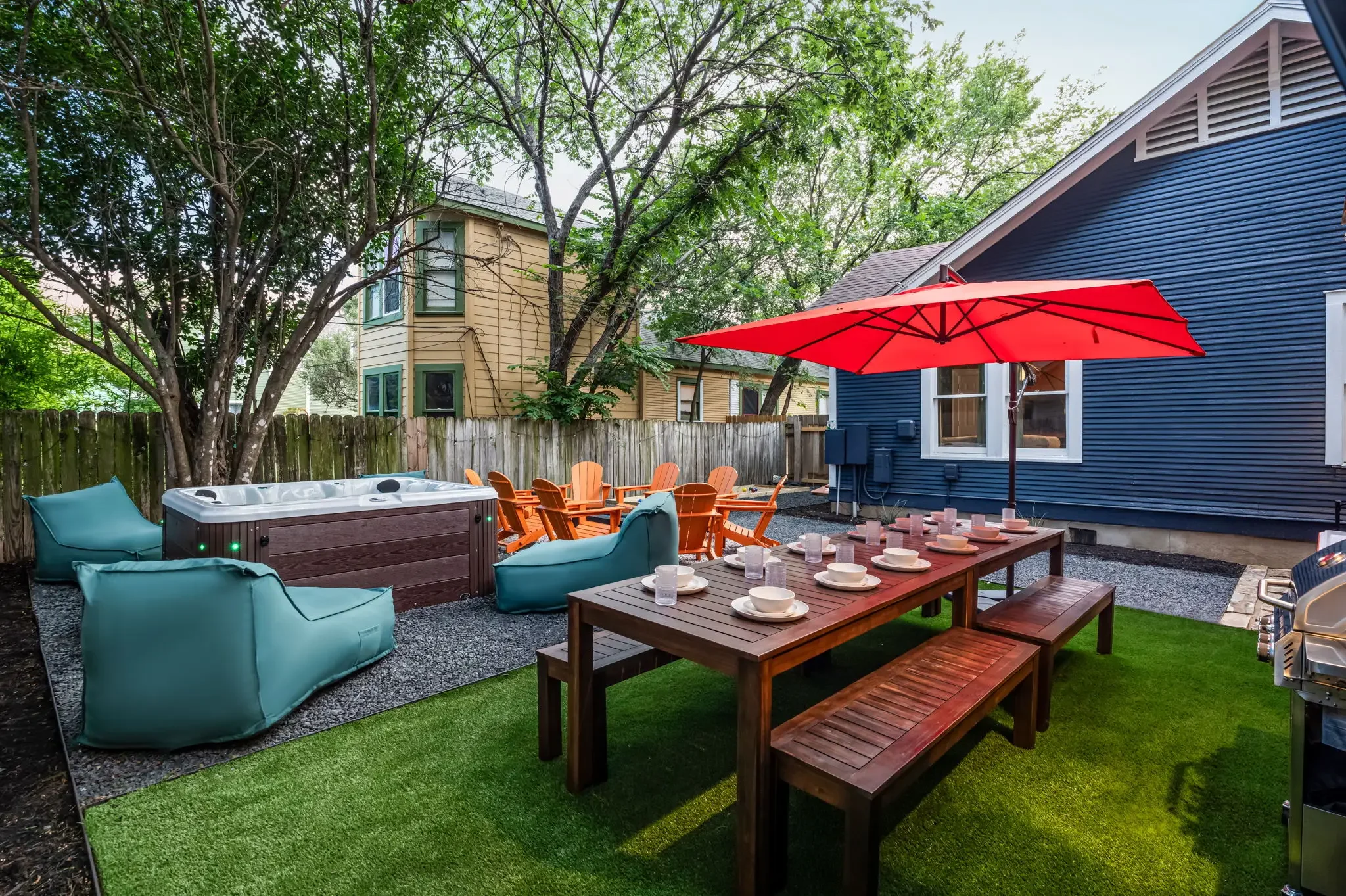 Airbnb backyard design with hot tub, long wood dining table, teal bean bags, orange Adirondack chairs, and red umbrella — Austin TX.