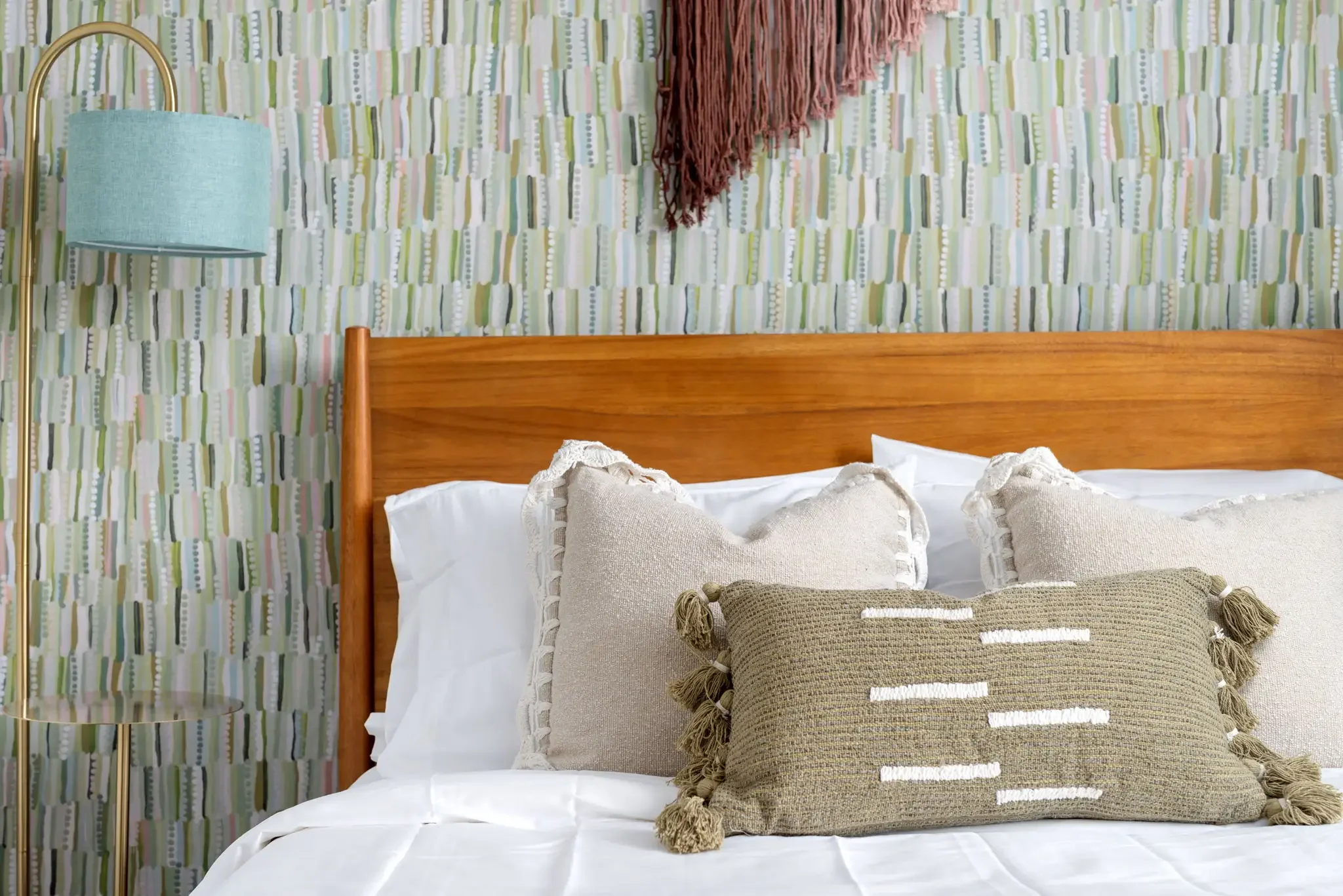 Short-term rental bedroom styling with green abstract wallpaper, wood headboard, teal lamp, and tassel pillow — Austin TX.