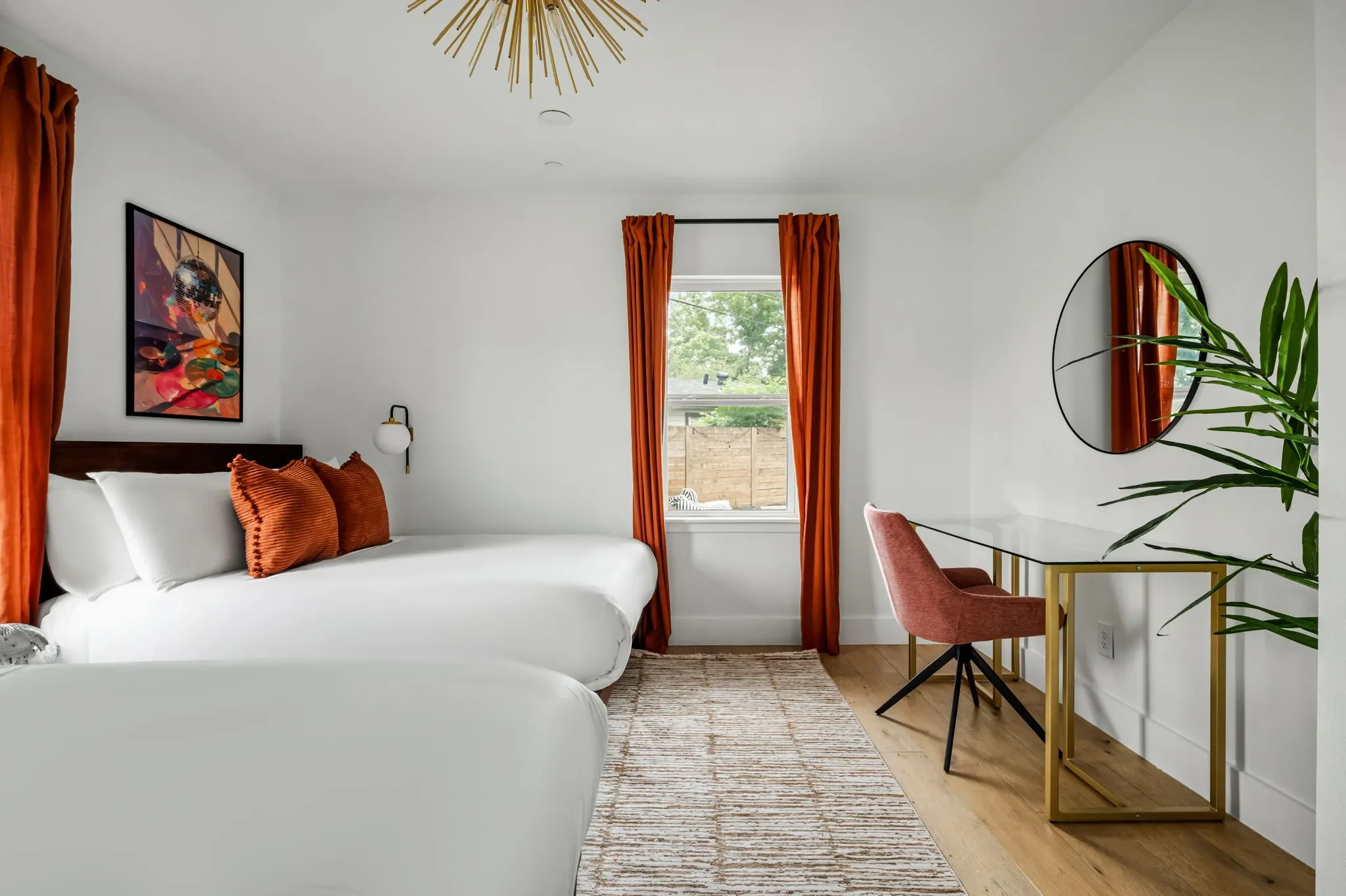 Airbnb bedroom design with queen bed, rust orange curtains, round mirror, pink accent chair, and gold desk — Austin TX.
