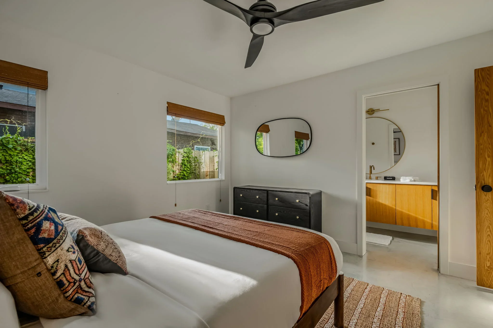 Short-term rental guest bedroom design with earth tone pillows, black dresser, oval mirror, and view into ensuite bathroom — Austin TX.