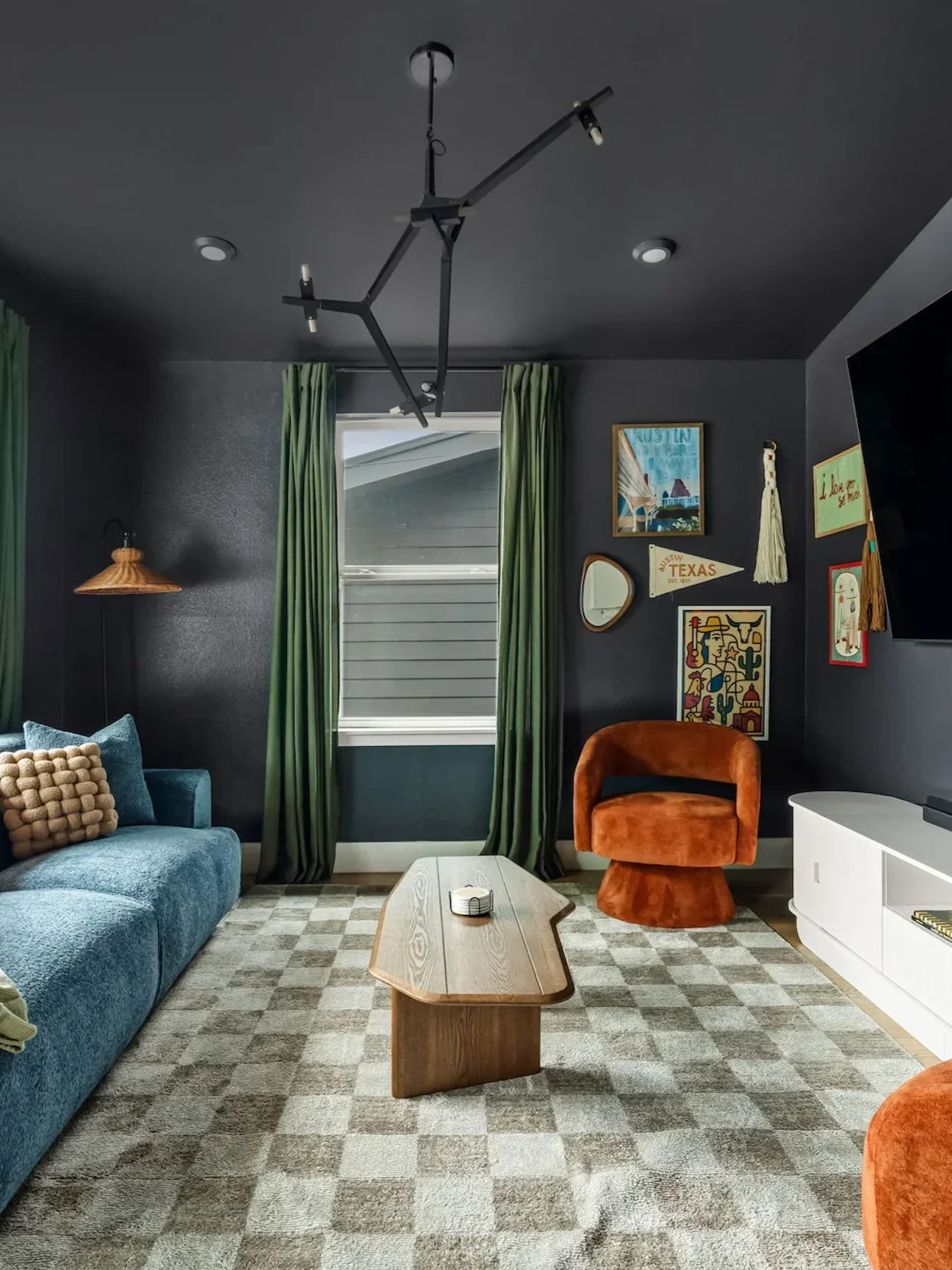 Short-term rental living room design with dark walls, blue velvet sofa, orange accent chair, and checkered rug — House of Ellie Interiors Austin TX.