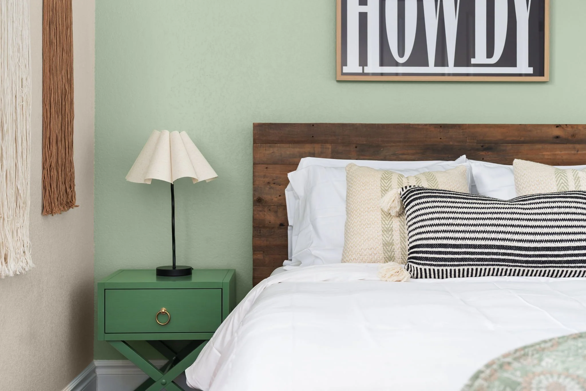 Short-term rental bedroom design with sage green walls, reclaimed wood headboard, green nightstand, and Howdy art — Austin TX.