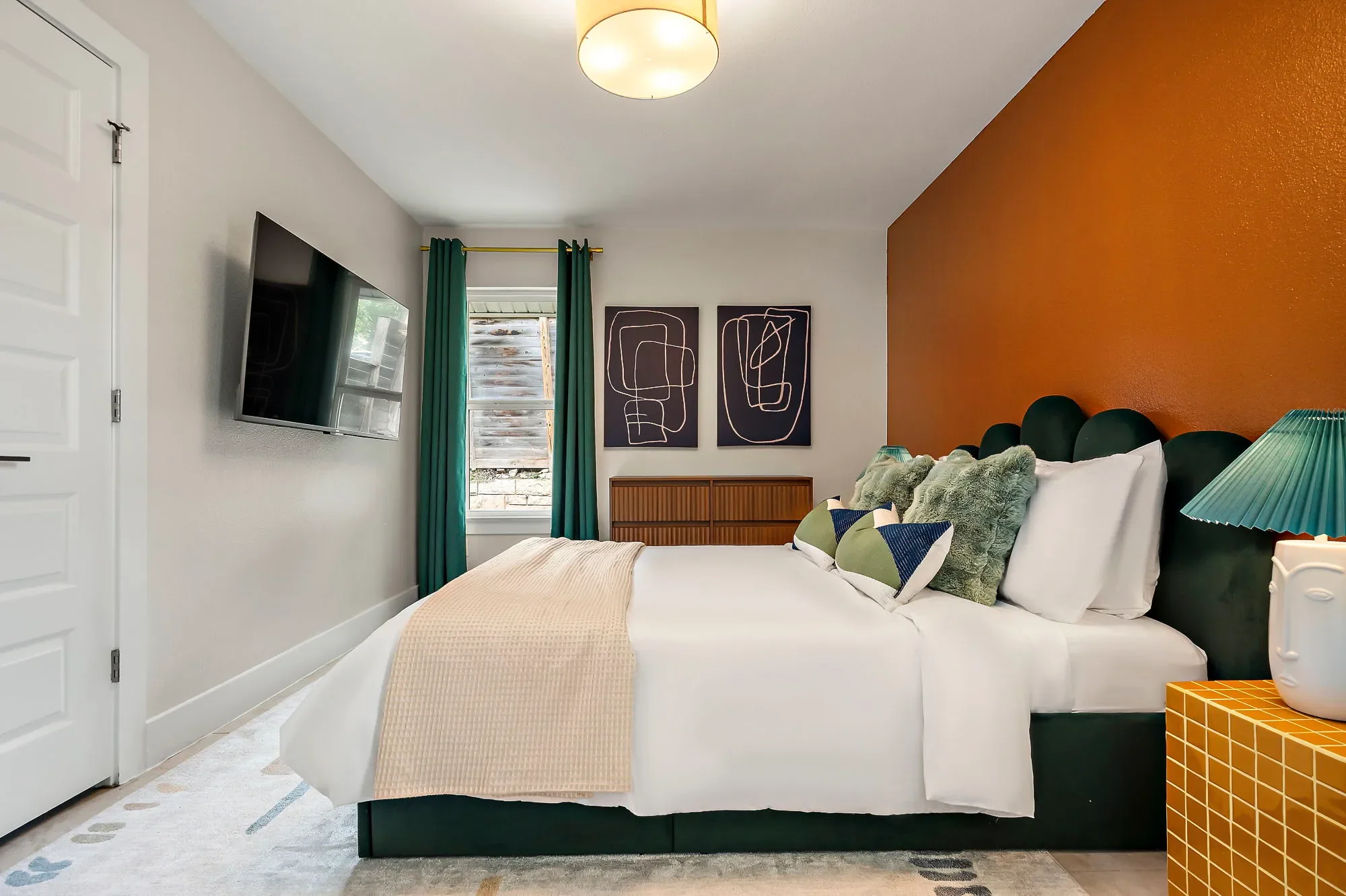 Bedroom featuring bold orange walls, green velvet bed, matching green curtains, navy wall art, and sculptural lamps on nightstands.