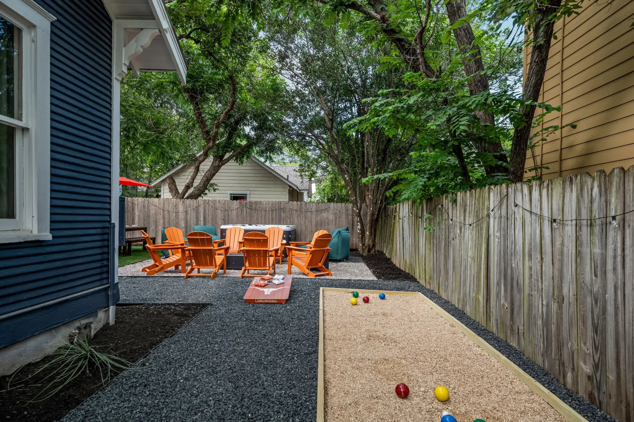 Airbnb backyard design with bocce ball court, orange Adirondack chairs, gravel patio, and string lights — Austin TX.
