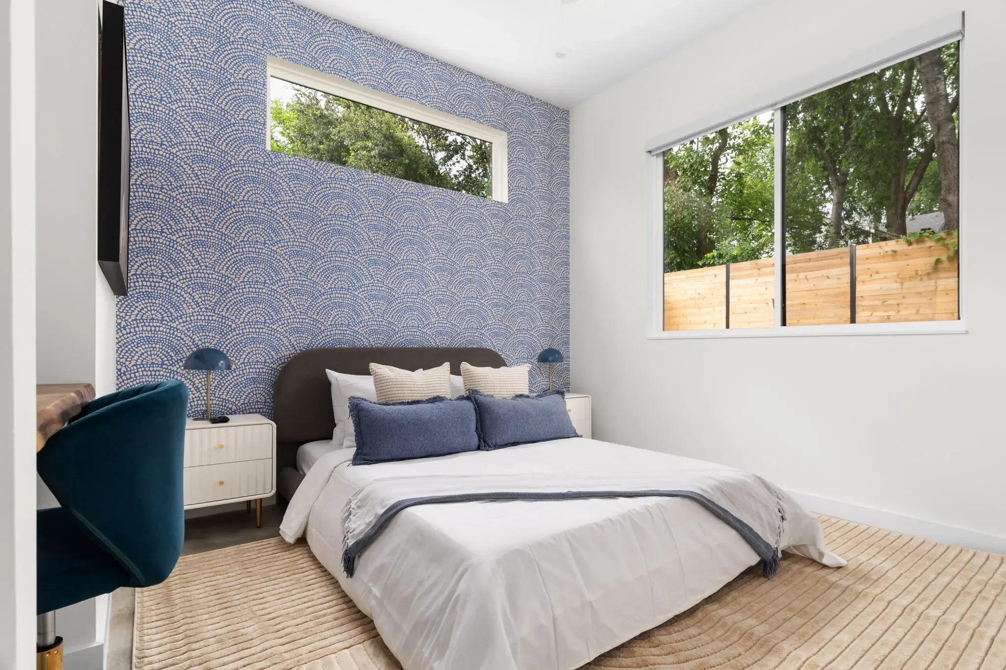 Airbnb bedroom design with blue scallop wallpaper, dark upholstered headboard, teal accent chair, white nightstand, and vaulted ceiling — Austin TX.
