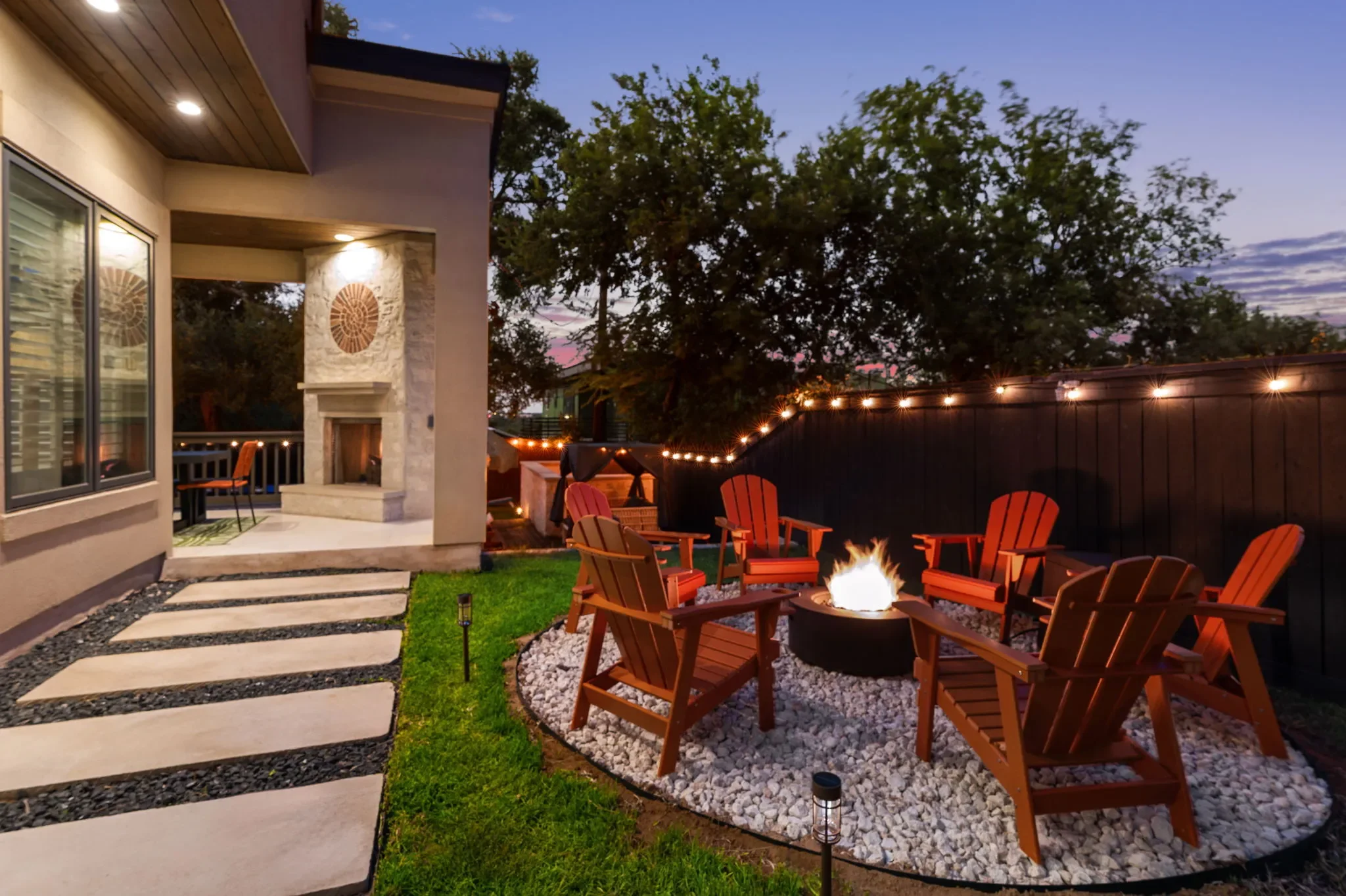 Short-term rental backyard design with fire pit, red Adirondack chairs, string lights, and stone fireplace at dusk — Austin TX.