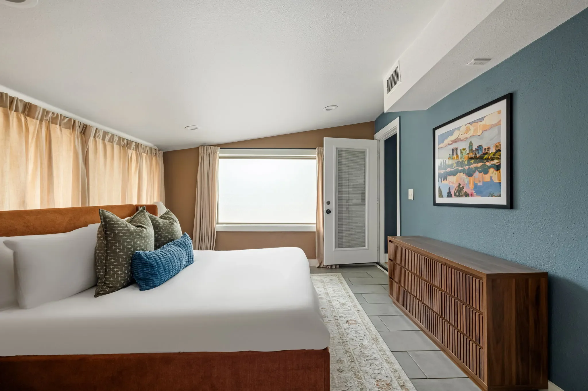 Airbnb bedroom design detail with blue accent wall, wood dresser, linen curtains, colorful city artwork, and layered pillows — Austin TX.