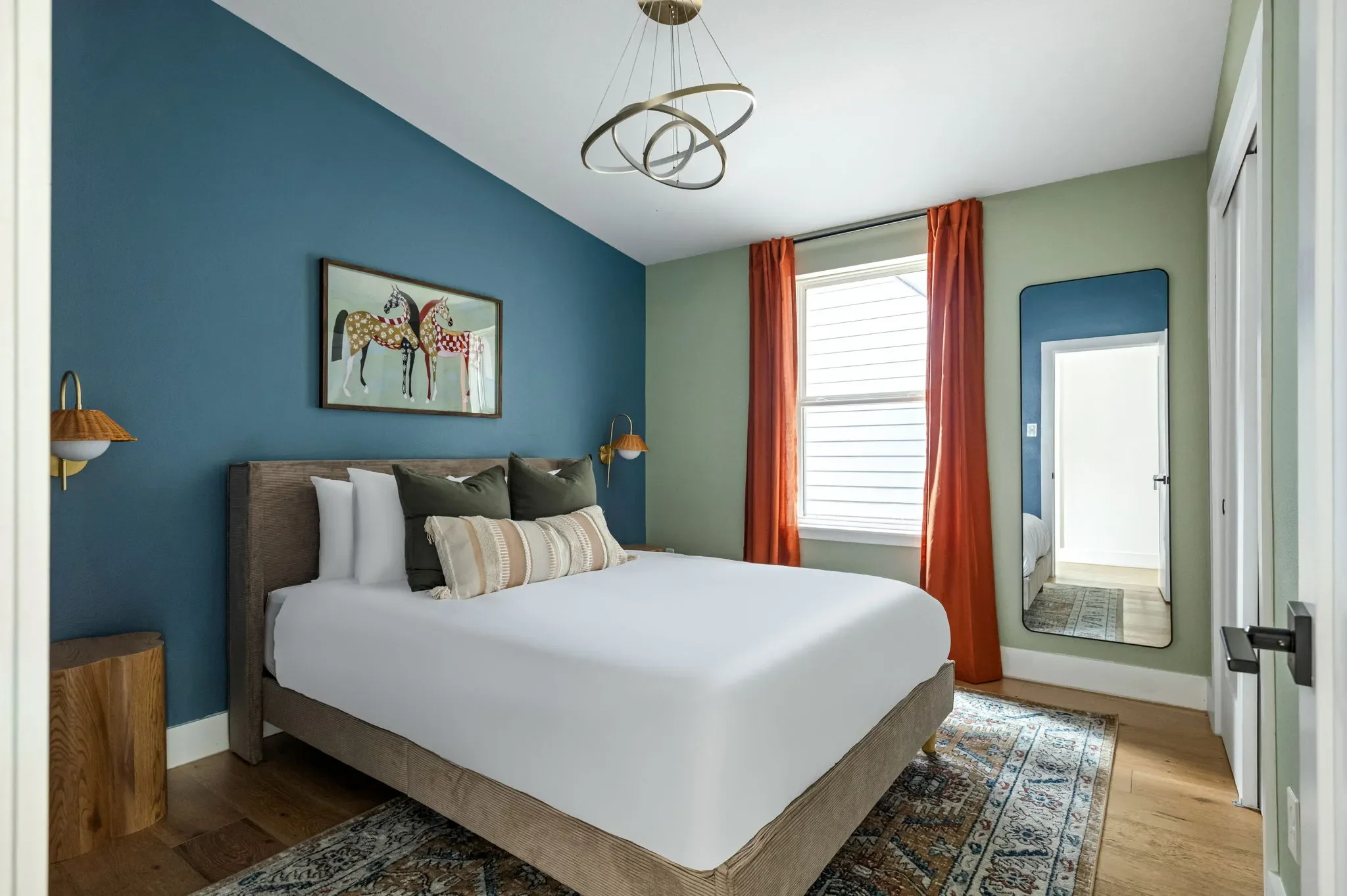 Airbnb bedroom design with blue accent wall, wood headboard, horse artwork, gold sconces, and orange curtains — Austin TX.
