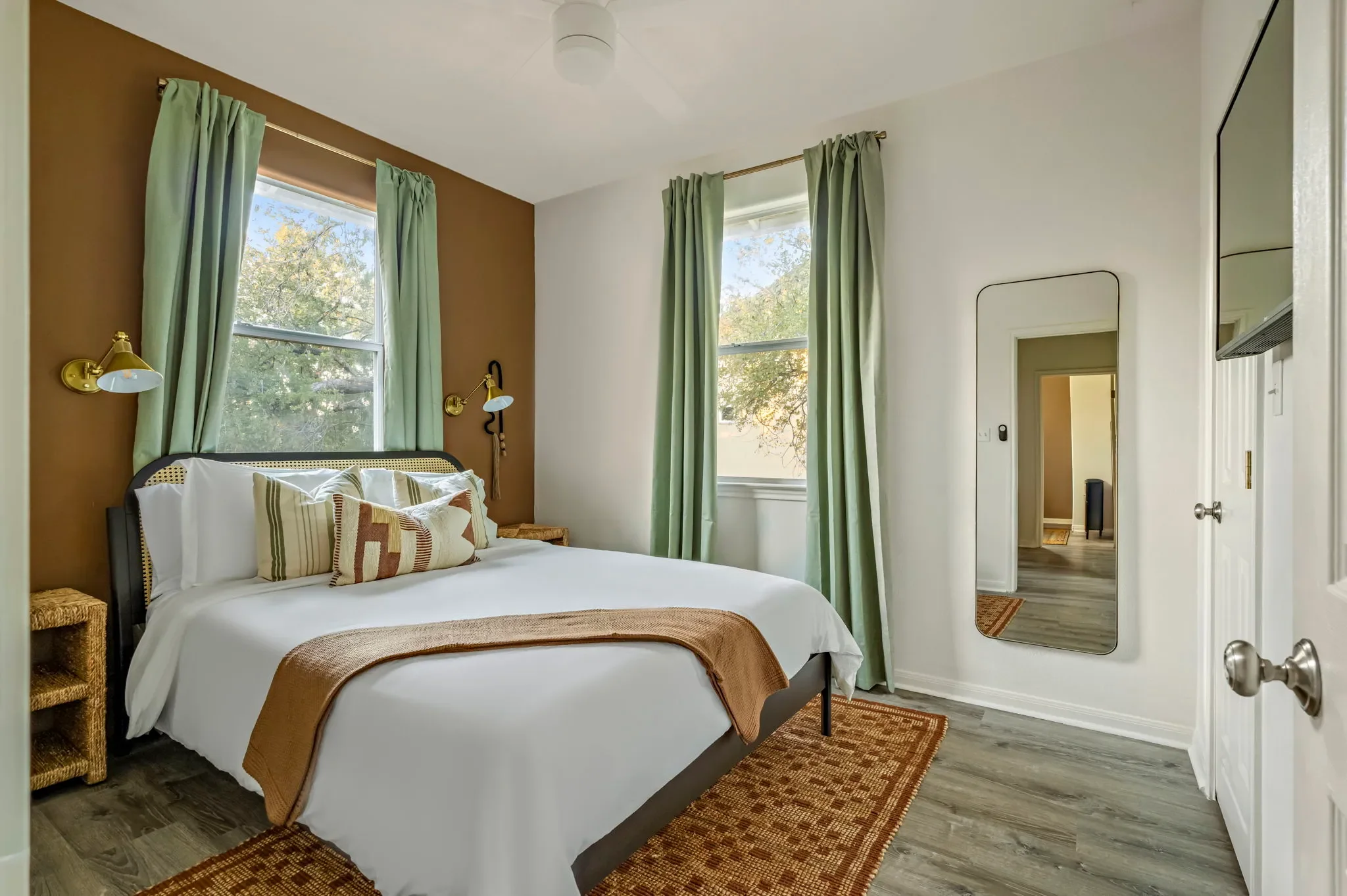 Bedroom with a black rattan bed frame, rattan nightstand, sage green floor-to-ceiling curtains, western-style area rug, beige walls, and a full-length mirror.