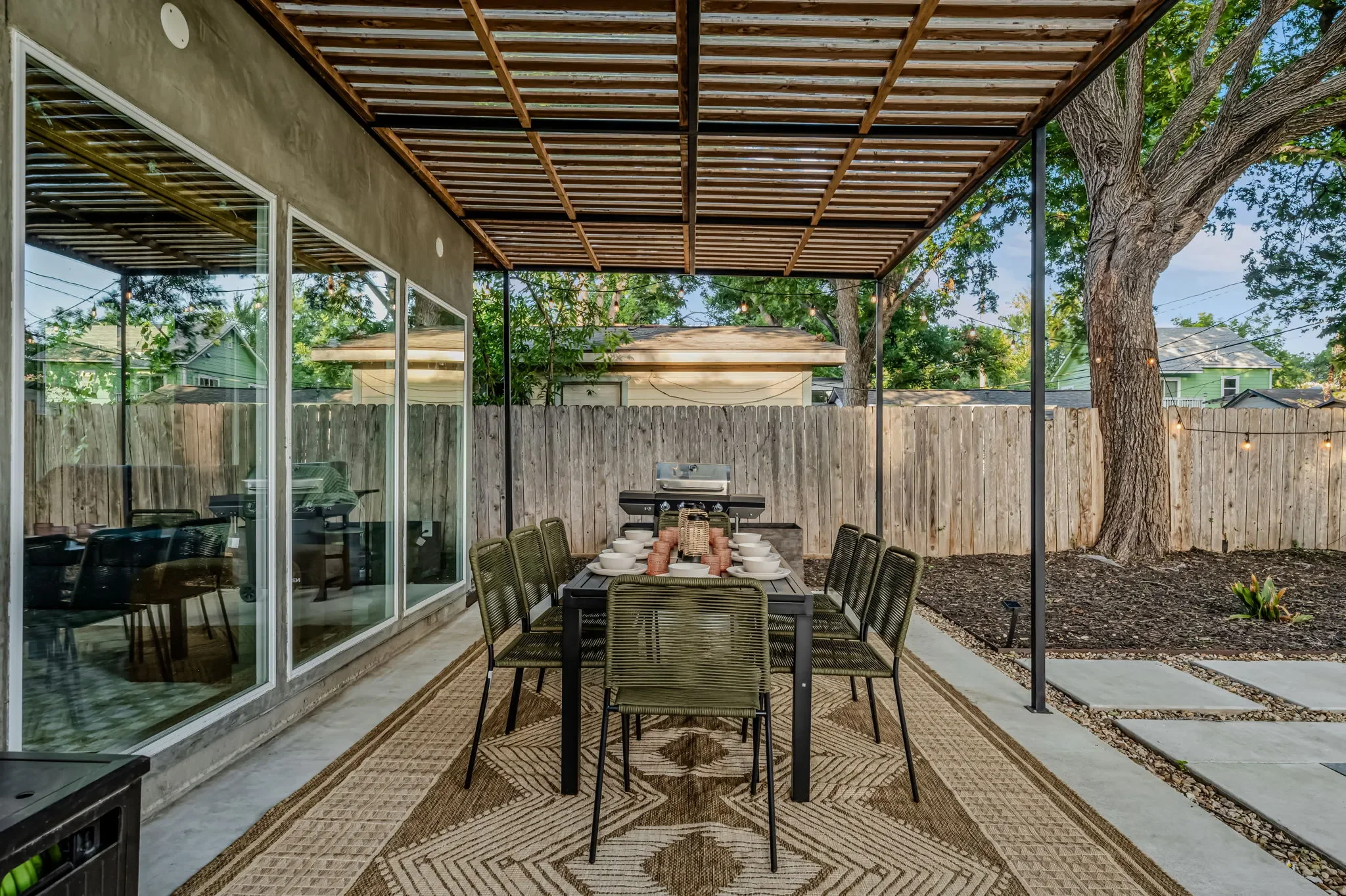Airbnb outdoor dining design with steel pergola, long dining table set for guests, and view to mature oak tree — Austin TX.
