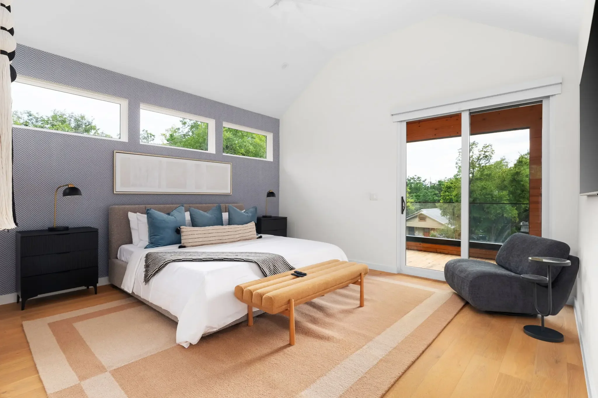 Short-term rental bedroom design with blue herringbone wallpaper, upholstered headboard, wood bench, accent chair, and sliding glass door — Austin TX.