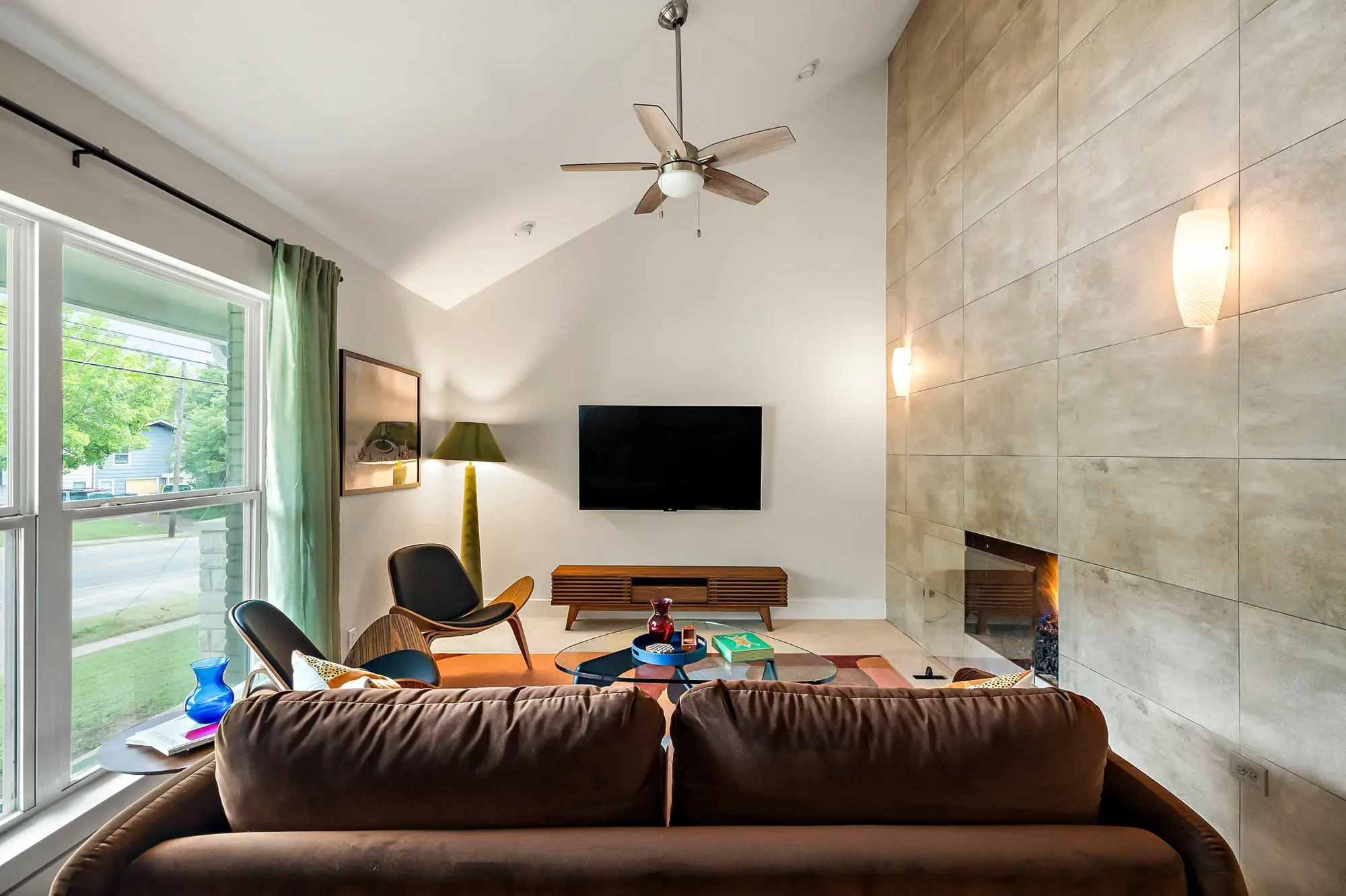Living room featuring a brown velvet sofa, mid-century modern lounge chairs, green velvet floor lamp, mounted TV, and floor-to-ceiling green curtains.