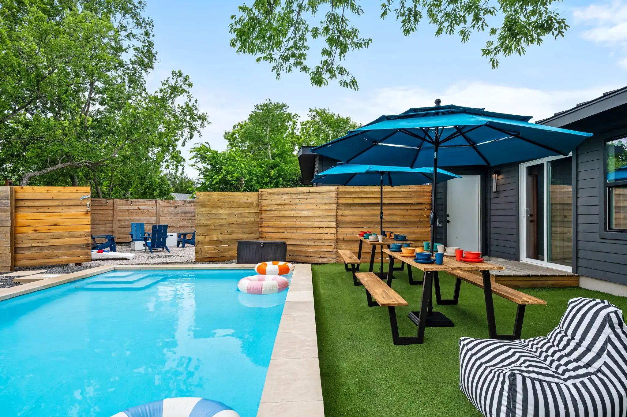Short-term rental backyard design with pool, picnic tables, blue umbrella, striped bean bags, and fenced yard — Austin TX.