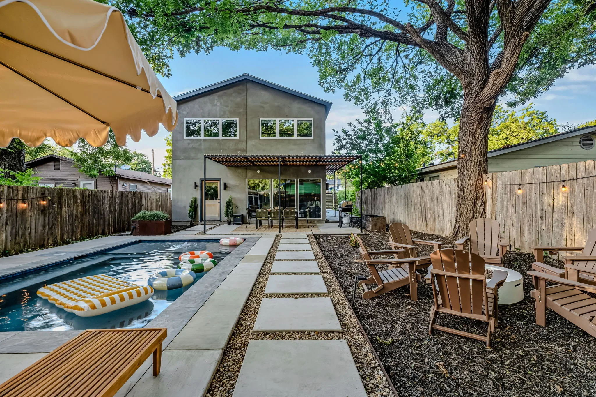 Airbnb backyard design with lap pool, scalloped umbrella, wood Adirondack chairs, pool float, and mature oak tree — Austin TX.
