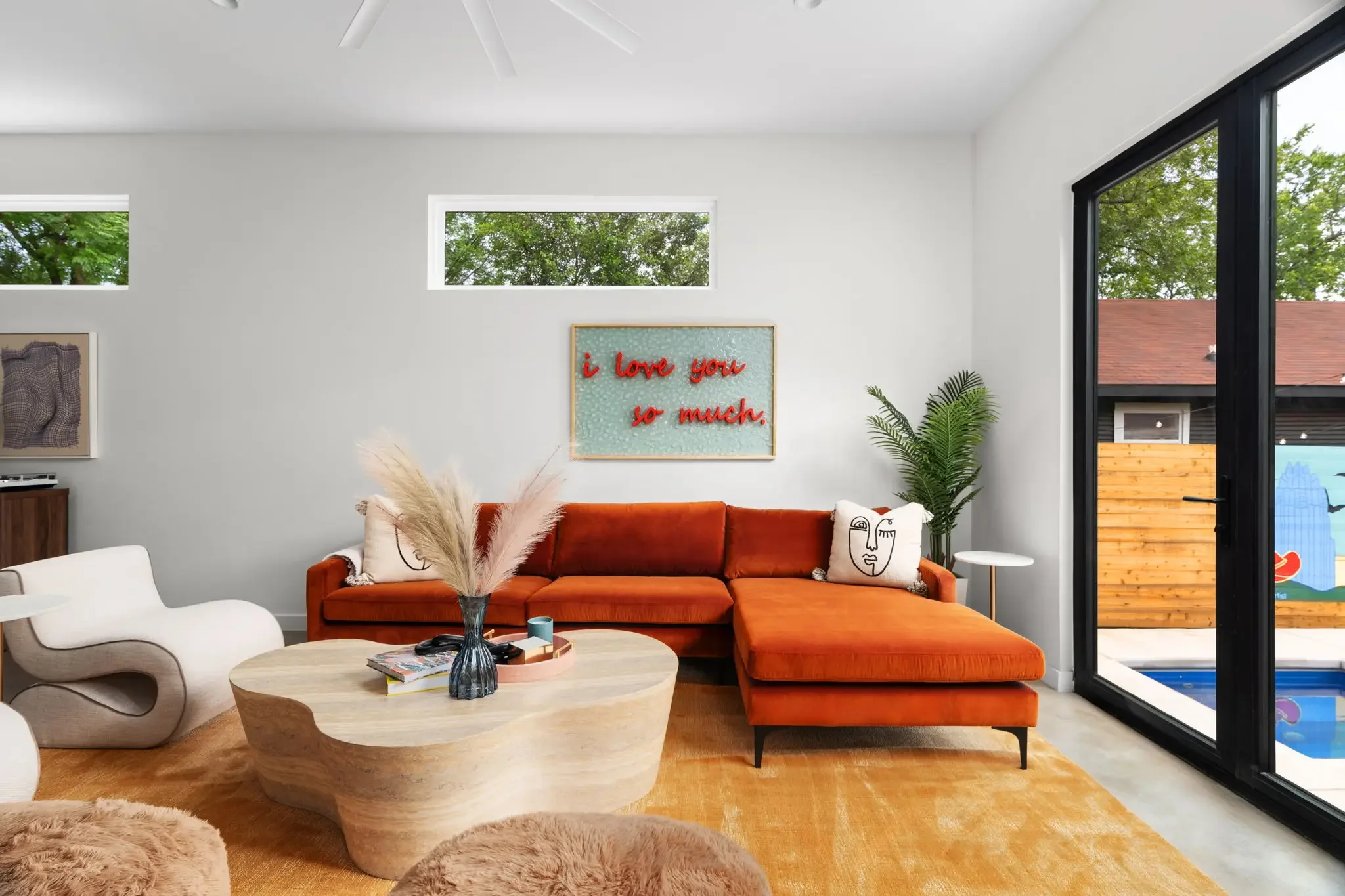 Short-term rental living room design with orange velvet sectional, organic coffee table, I Love You So Much neon art, and pool view — Austin TX.