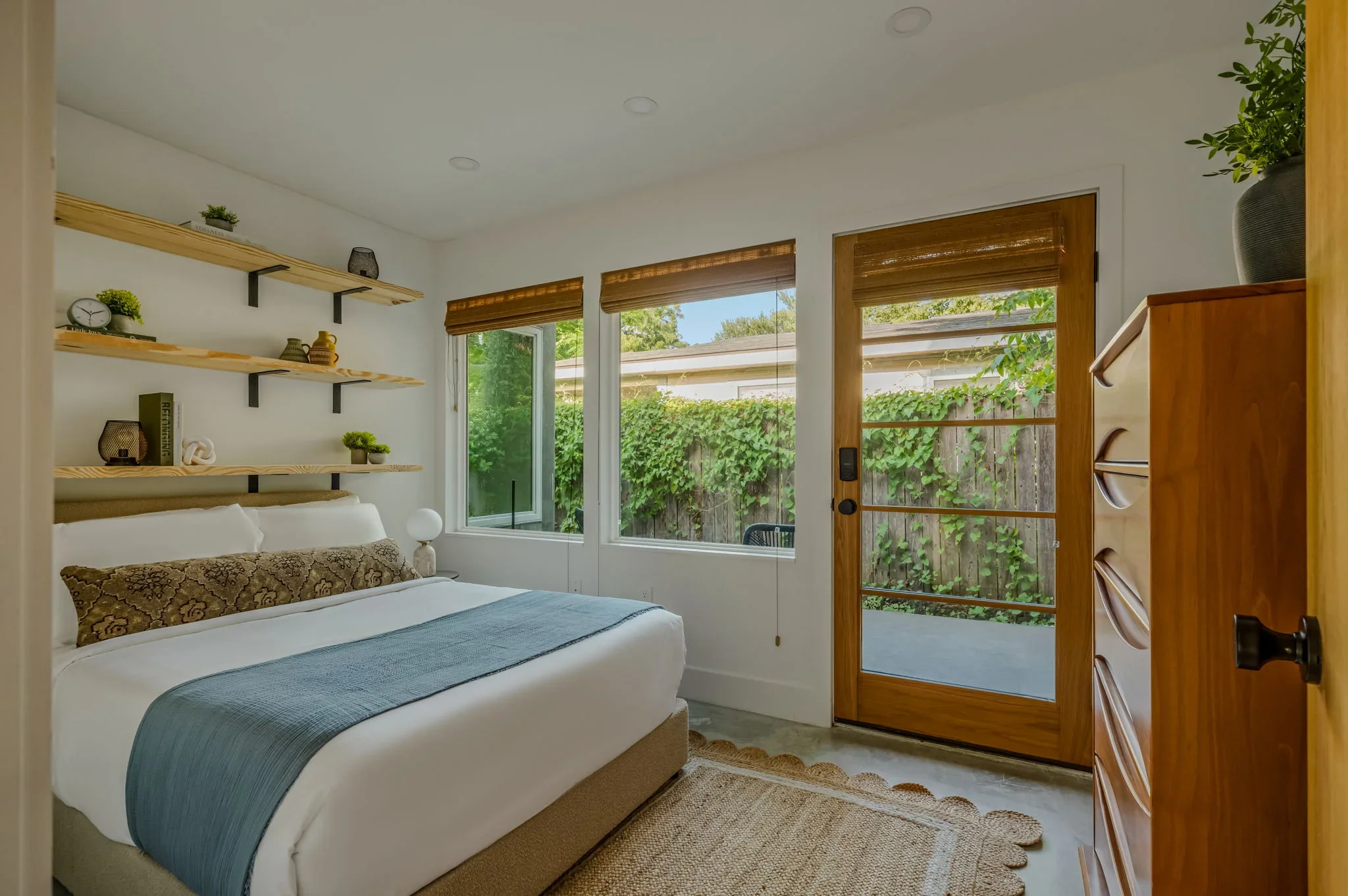 Short-term rental bedroom design with floating shelves, wood door to garden, bamboo shades, and blue throw — Austin TX.