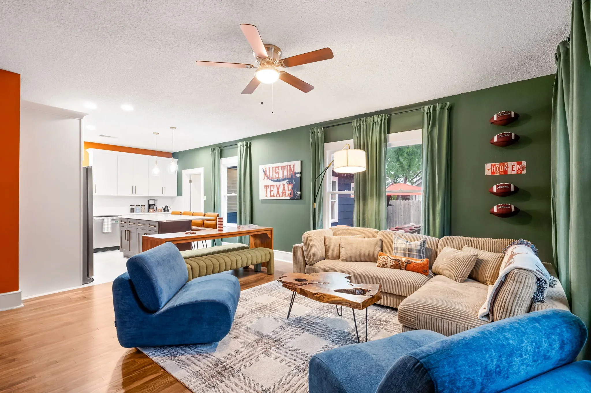 Short-term rental open plan design with dark green walls, tan sectional, blue accent chair, live edge coffee table, and white kitchen — Austin TX.