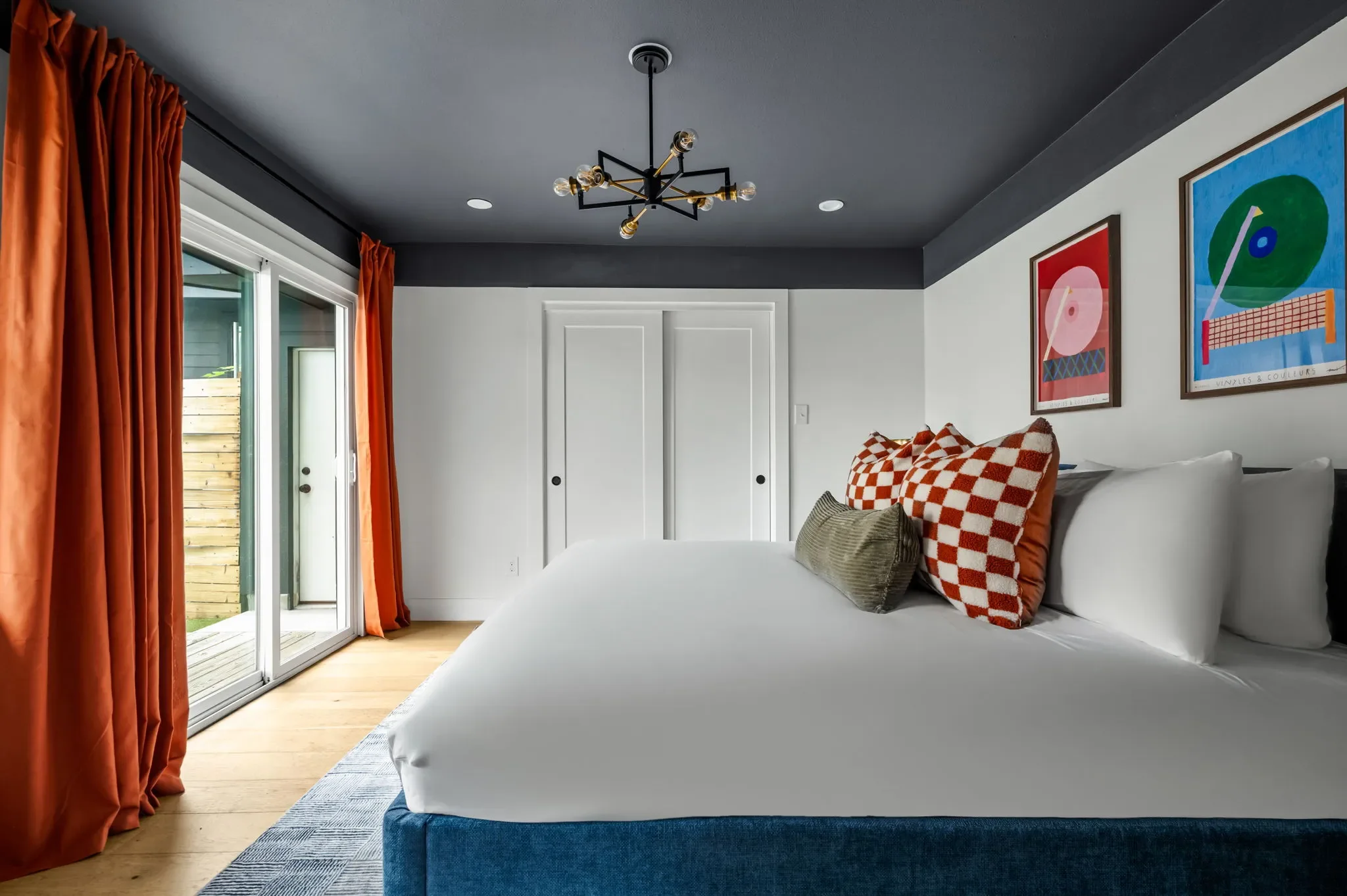 Airbnb guest house bedroom design with dark ceiling, blue upholstered bed, checkered pillows, orange curtains, and sliding glass door to backyard — Austin TX.