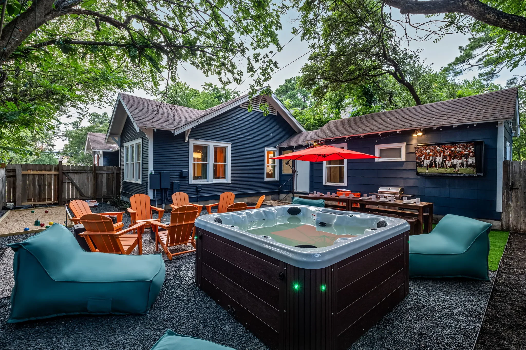 Short-term rental backyard design with hot tub, outdoor TV, orange Adirondack chairs, teal bean bags, and red umbrella — Austin TX.