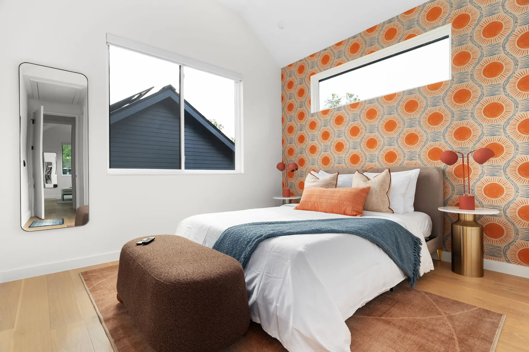 Bedroom with sun-style orange wallpaper, brown bench, taupe bed frame, orange table lamps, and a full-length mirror.