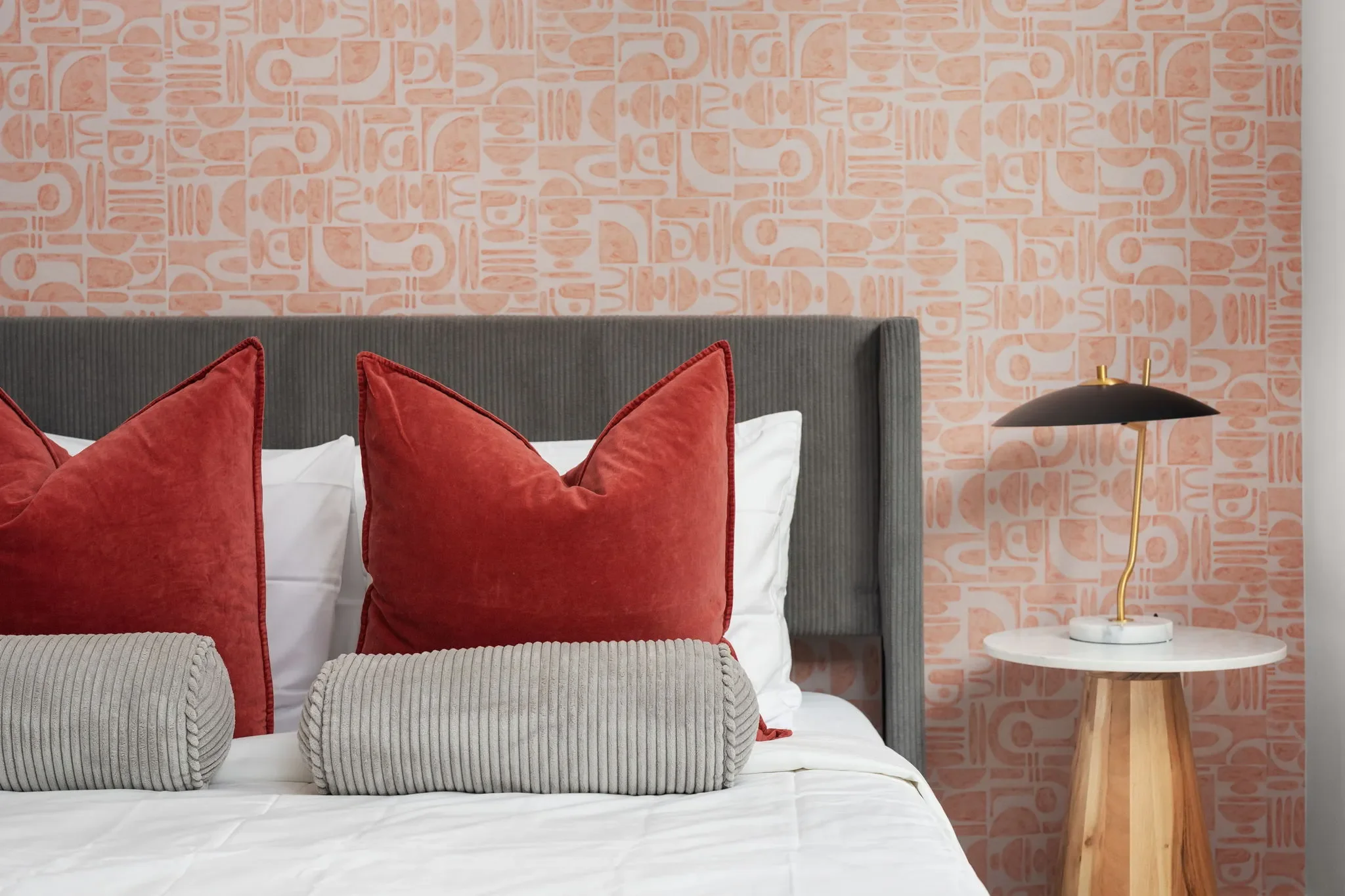Bedroom with geometric pink wallpaper, grey fabric bed frame, marble and wood nightstand with modern table lamps, and red decorative pillows.