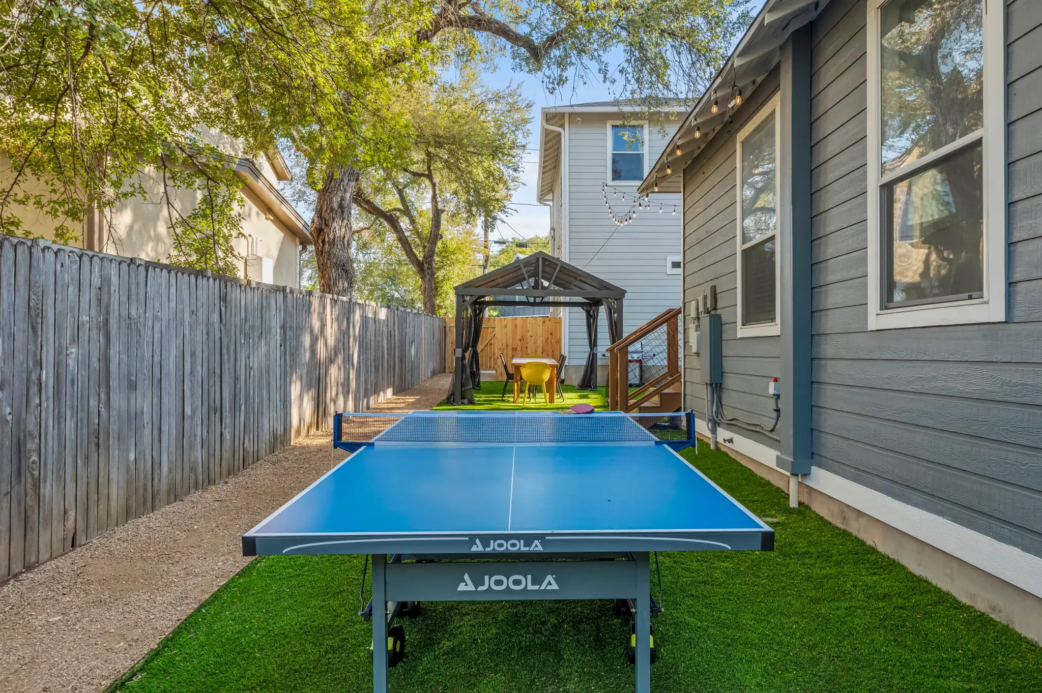 Backyard space featuring a table tennis table on artificial turf, with a gazebo dining area beyond.