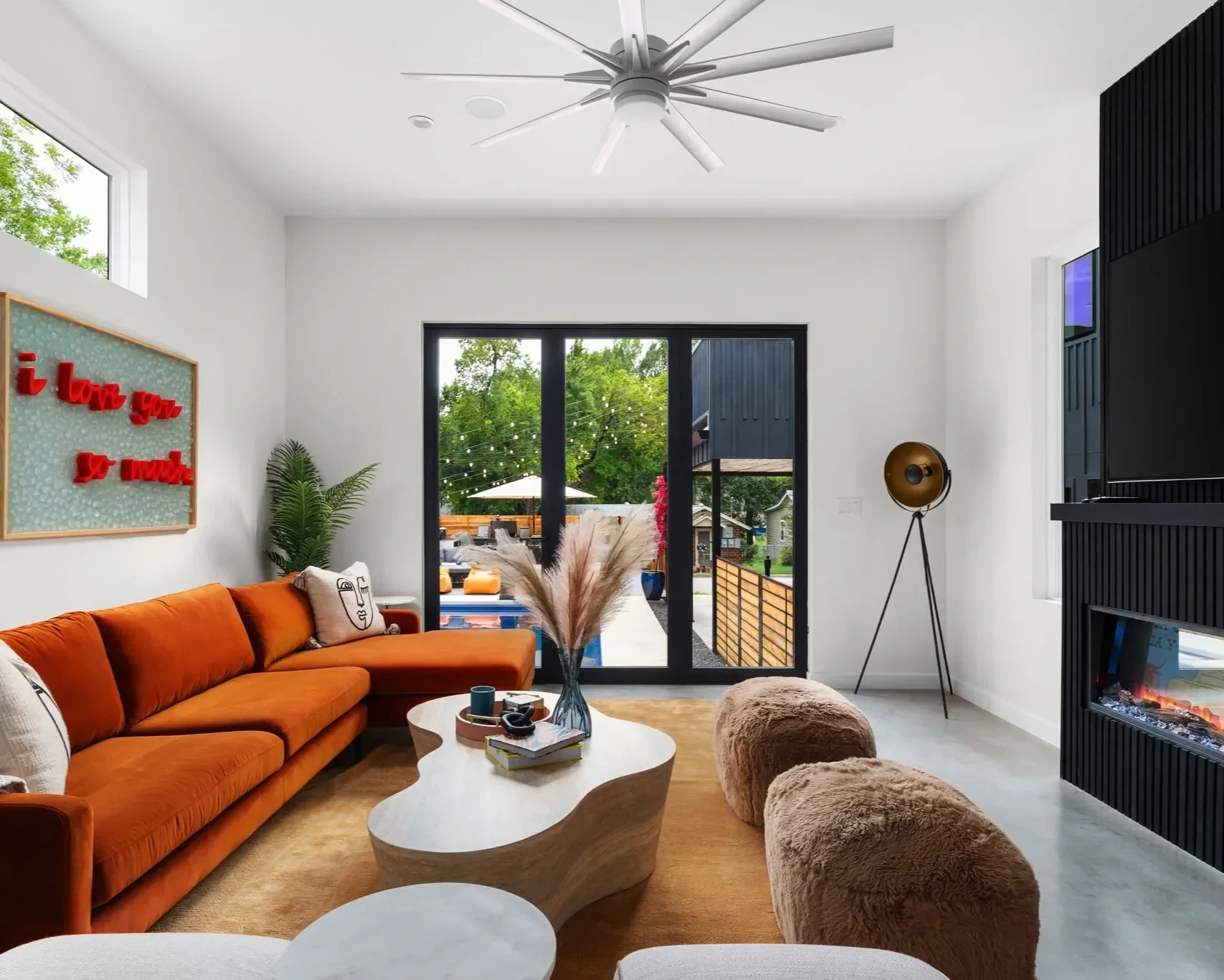 Modern short-term rental living room with rust-colored sectional sofa, sculptural coffee table, textured accent chairs, and large glass doors opening to an outdoor entertaining area.