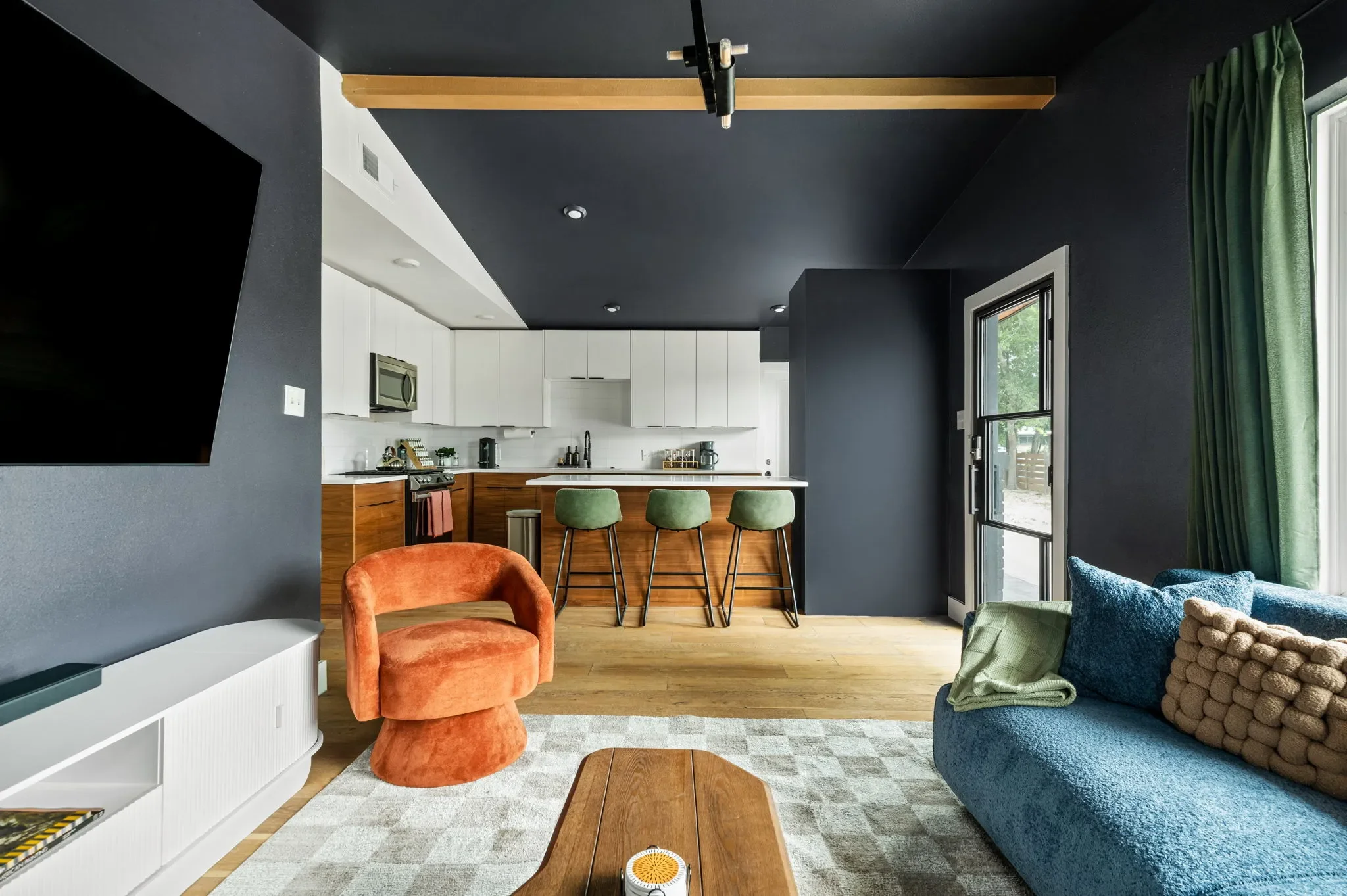 Airbnb open plan design with dark ceiling, white kitchen cabinets, wood island, green bar stools, and orange accent chair — Austin TX.