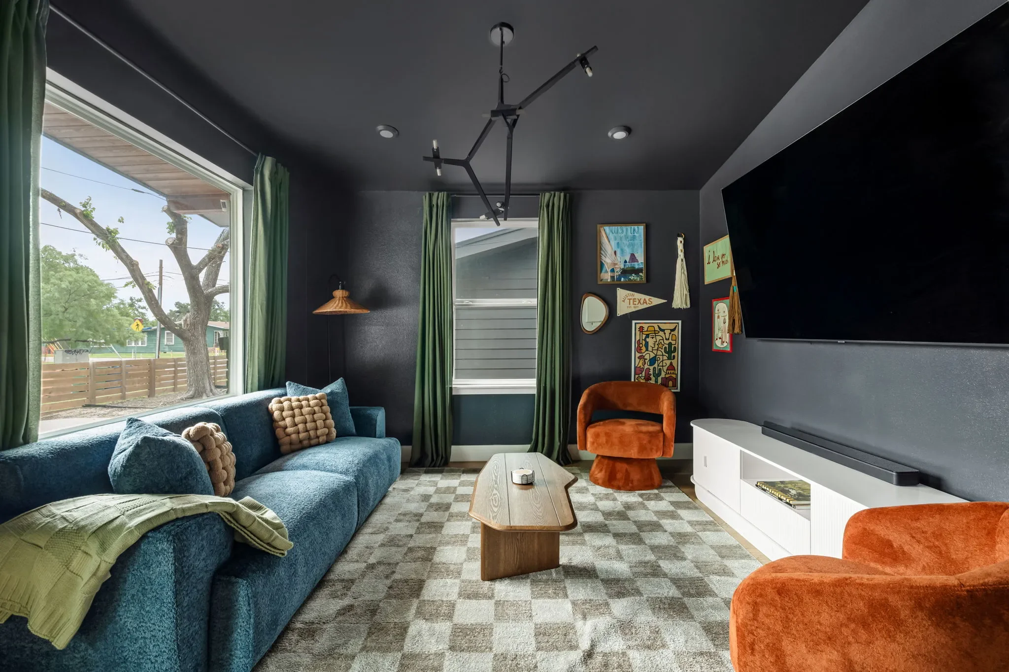 Short-term rental living room design with dark walls, blue sofa, orange accent chairs, checkered rug, and large TV — Austin TX.