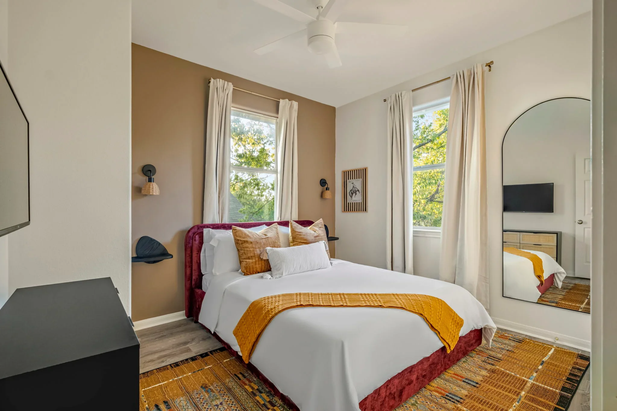 Short-term rental bedroom design with burgundy velvet headboard, tan accent wall, mustard throw, and arched mirror — Austin TX.