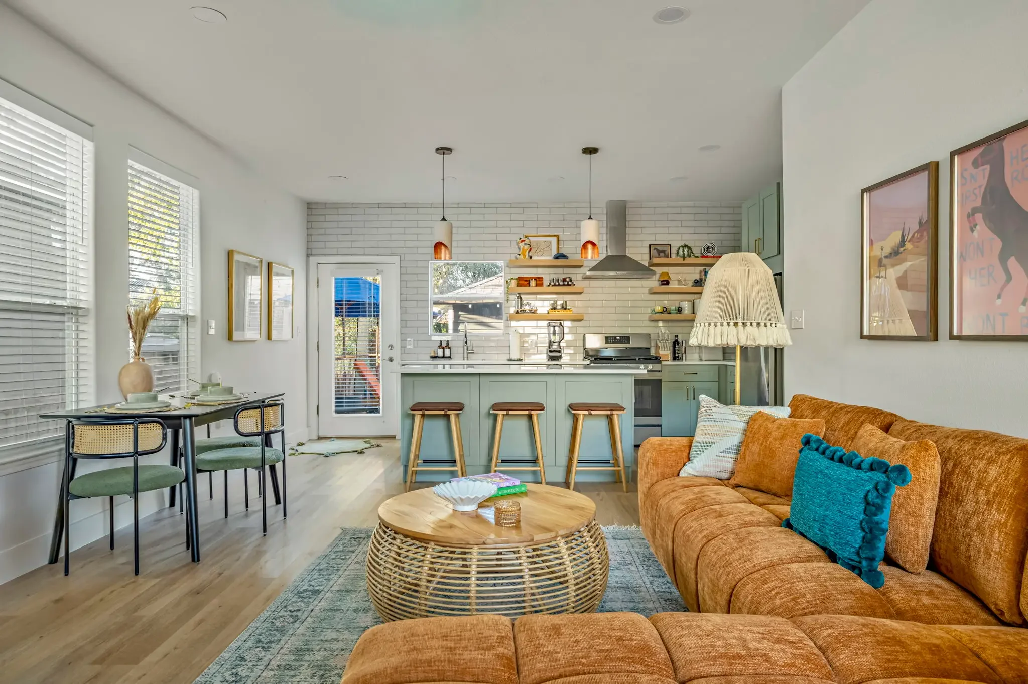 Short-term rental interior design with sage green kitchen, tan sectional sofa, teal pillows, and colorful wall art.