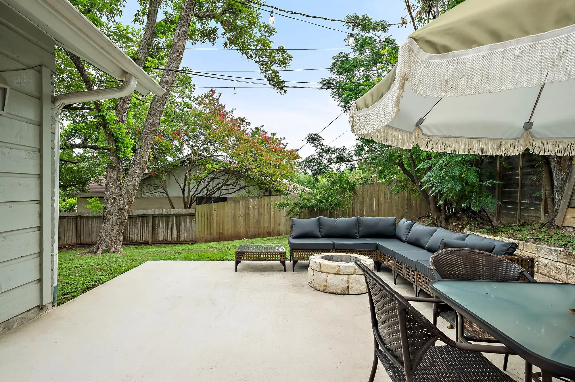 Short-term rental backyard design with concrete patio, dark sectional, macrame umbrella, fire pit, and mature trees — Austin TX.