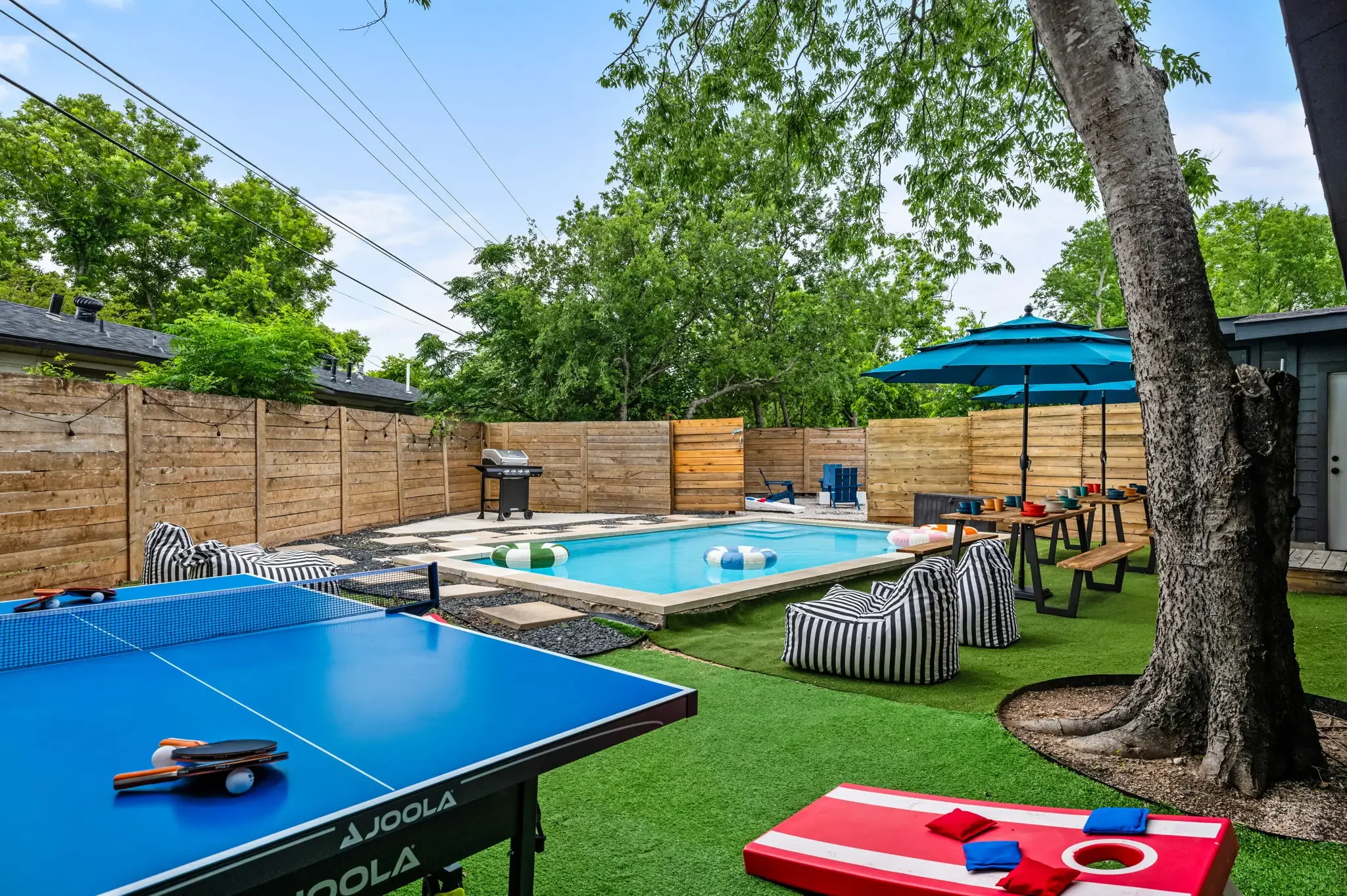 Airbnb backyard design with pool, ping pong table, cornhole, striped bean bags, and blue umbrella — Austin TX.