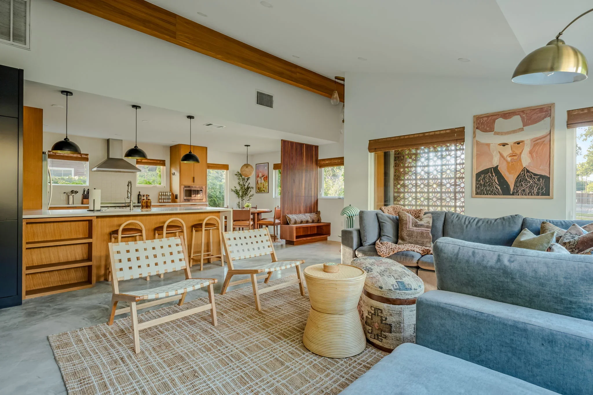 Short-term rental open plan design with blue sectional, wood beam ceiling, warm wood kitchen island, woven accent chairs, and cowboy portrait art — Austin TX.