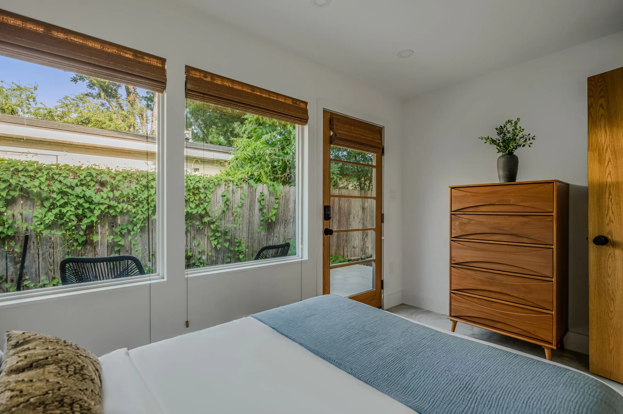 Airbnb bedroom design with large garden-view windows, mid-century wood dresser, and bamboo shades — Austin TX.