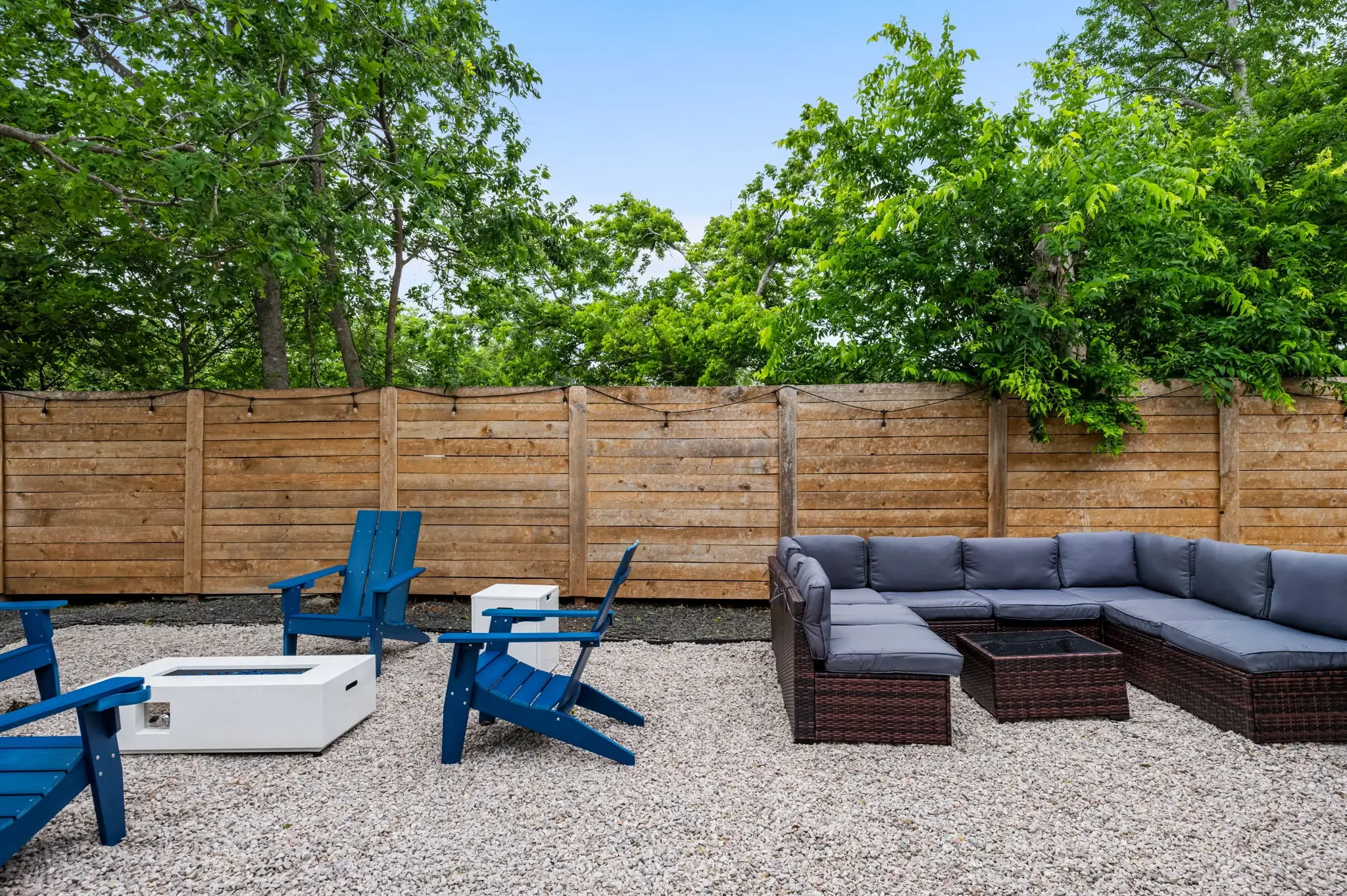 Airbnb backyard design with gravel patio, blue Adirondack chairs, fire pit, and large sectional seating under mature trees — Austin TX.