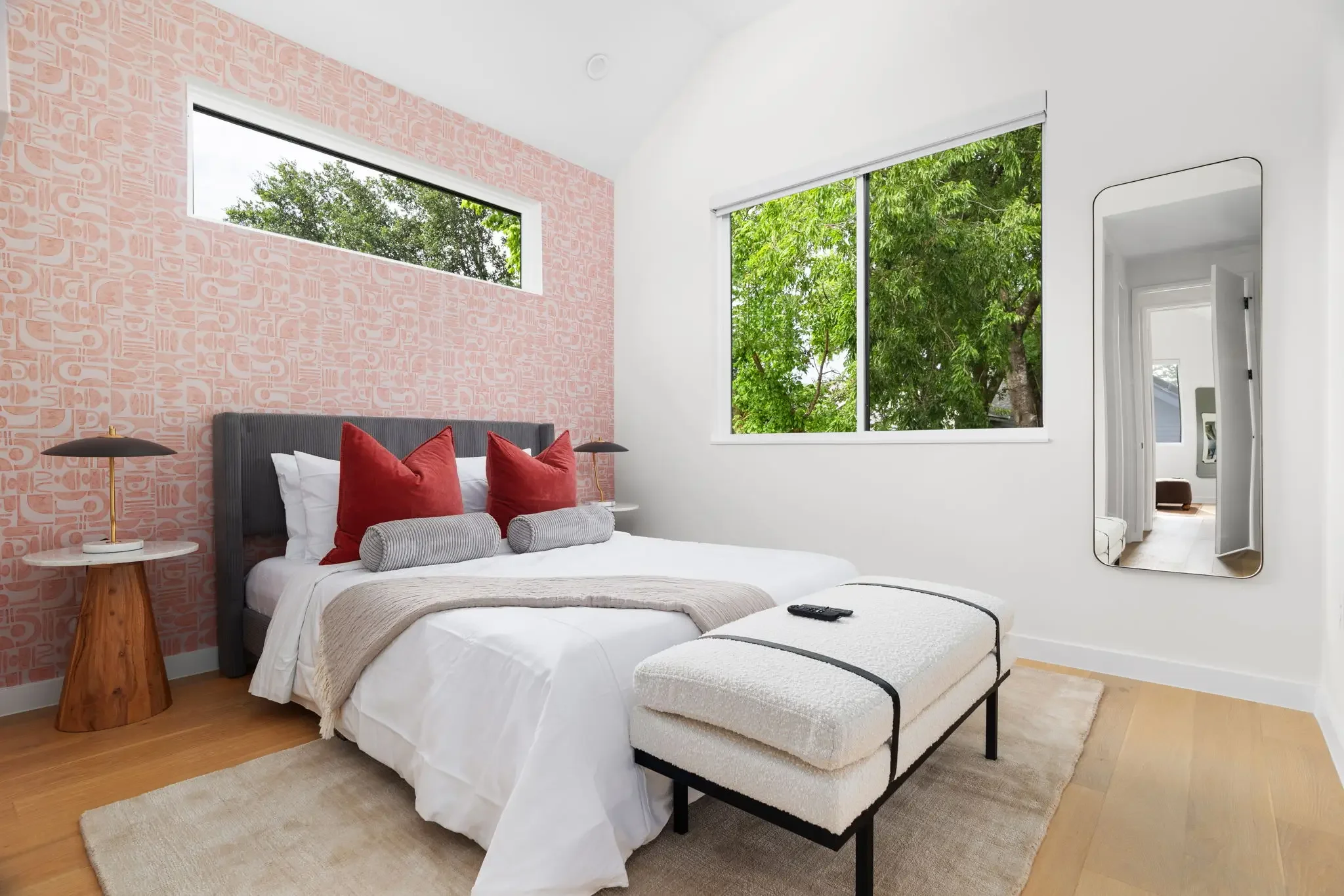Short-term rental bedroom design with pink abstract wallpaper, gray upholstered headboard, red pillows, wood nightstand, and leaning mirror — Austin TX.