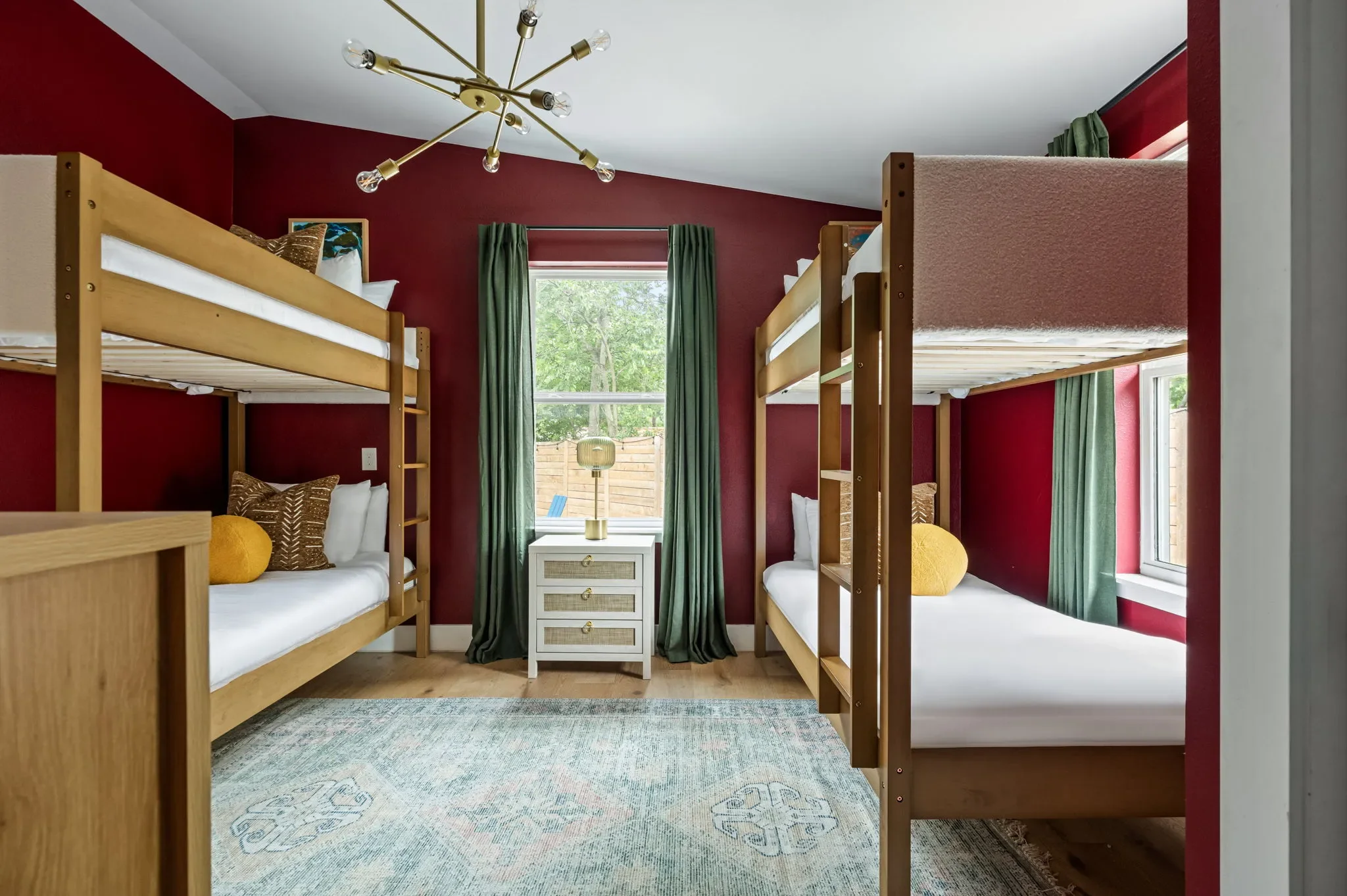 Airbnb bunk room design detail with red accent wall, wood bunk beds, yellow pillows, green curtains, and brass chandelier — Austin TX.