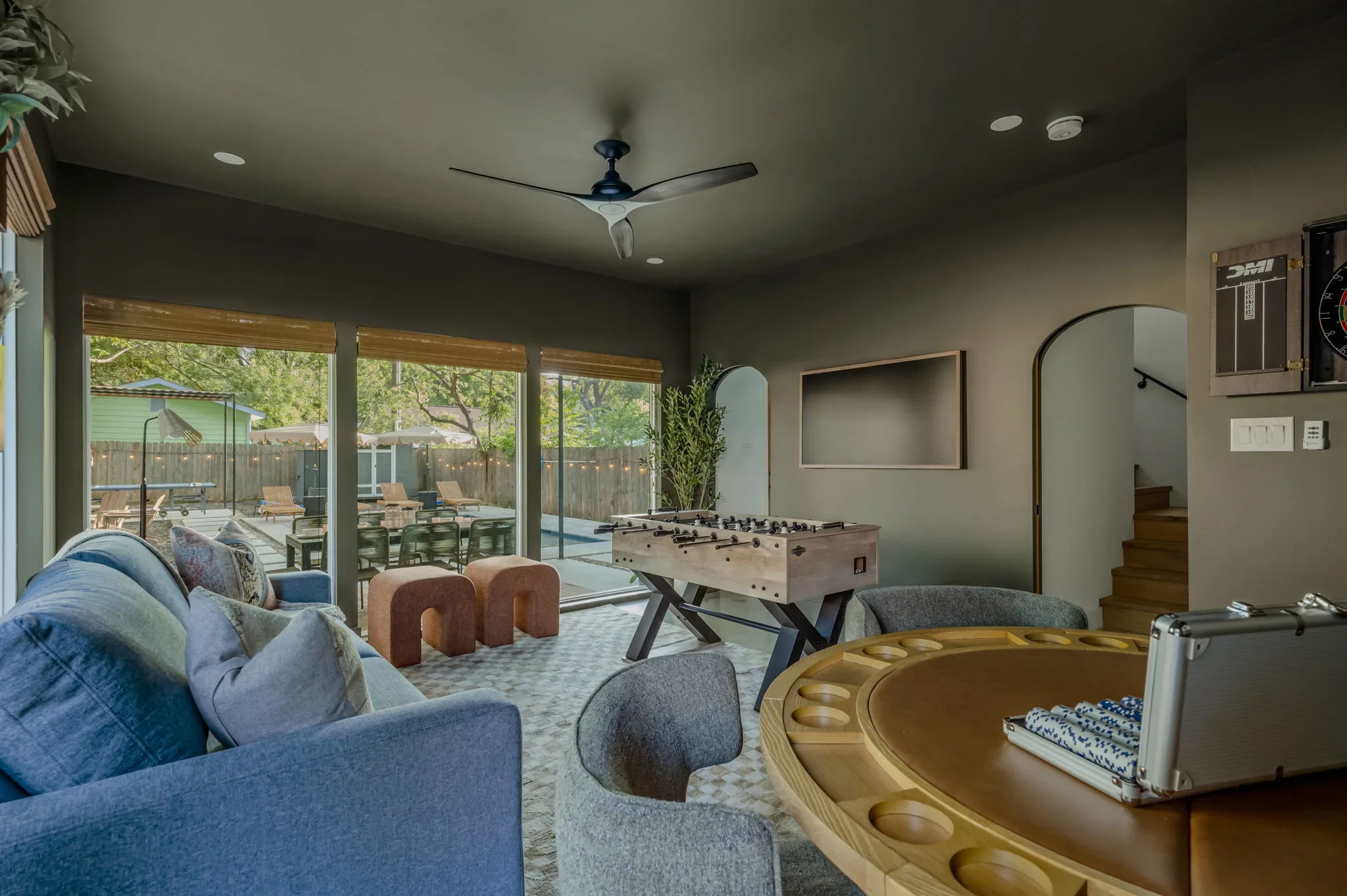 Short-term rental game room design with deep olive green walls, blue sofa, foosball table, arched doorway, and pool view — Austin TX.