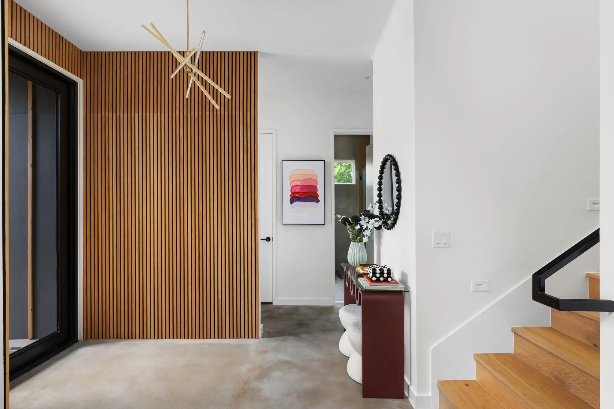Airbnb entryway design with floor-to-ceiling wood slat accent wall, gold cross chandelier, red console table, and open staircase — Austin TX.