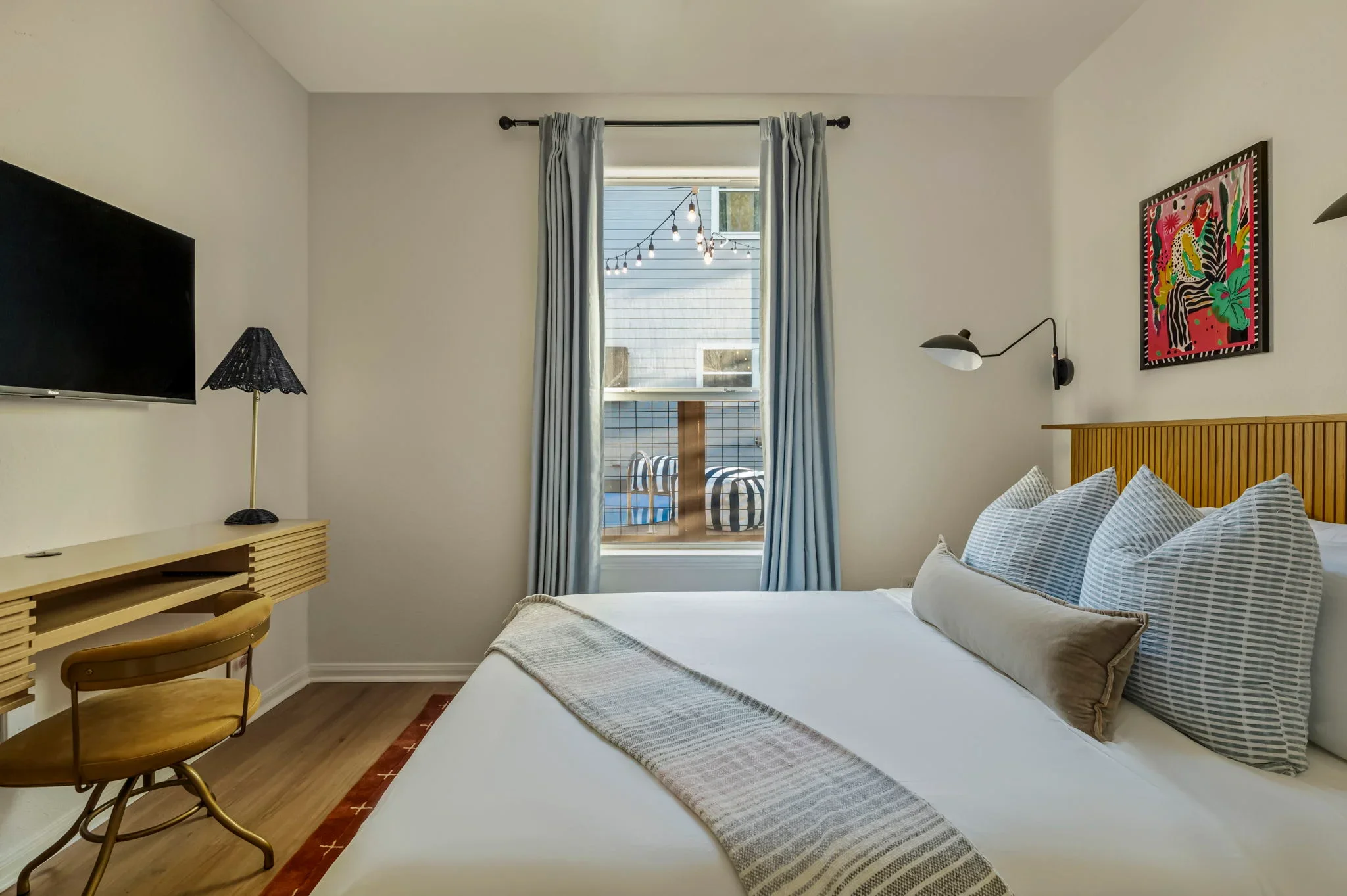 Airbnb bedroom design with wood slat headboard, blue curtains, TV, wood desk, and colorful figure wall art — Austin TX.