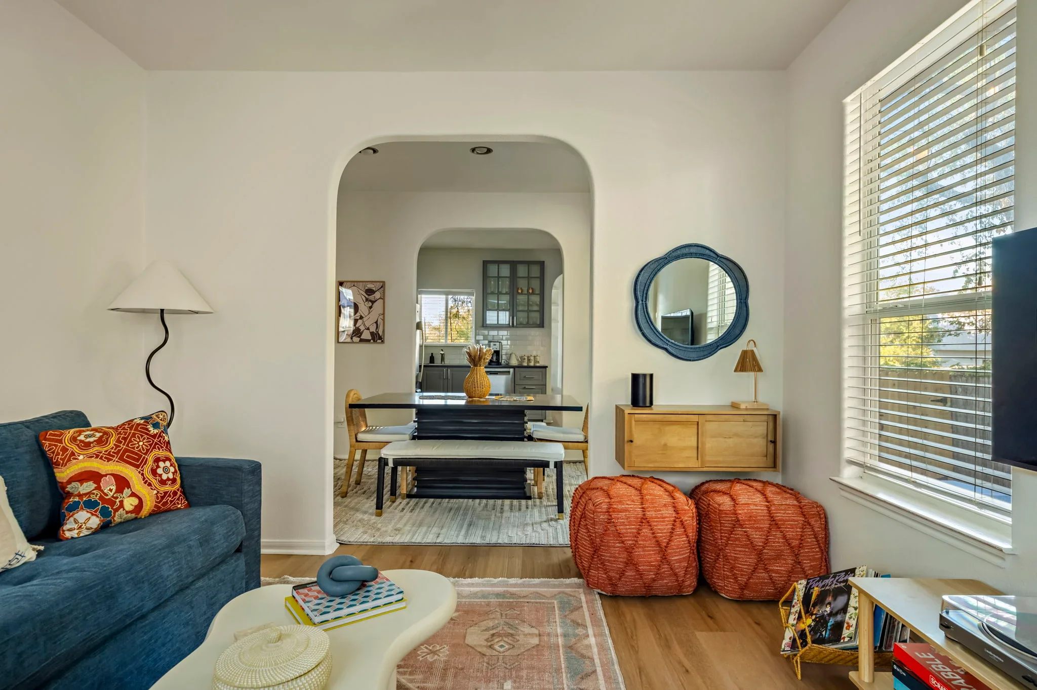 Airbnb living room design with navy sofa, rust pouf ottomans, scalloped blue mirror, and view through arched doorway to dining and kitchen — Austin TX.