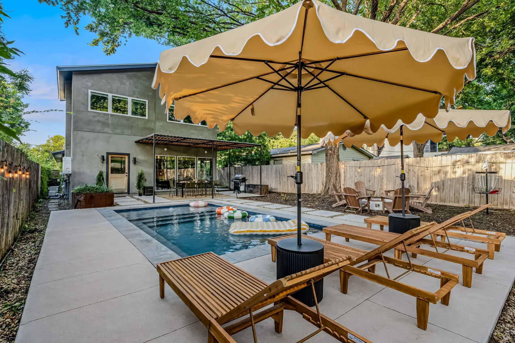 Short-term rental pool design with scalloped gold umbrella, wood lounge chairs, pool floats, and pergola — Austin TX.